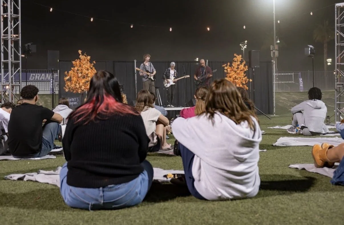 People watching a live outdoor concert at night, sitting on blankets on the grass, with a band performing on stage decorated with orange trees and string lights overhead.