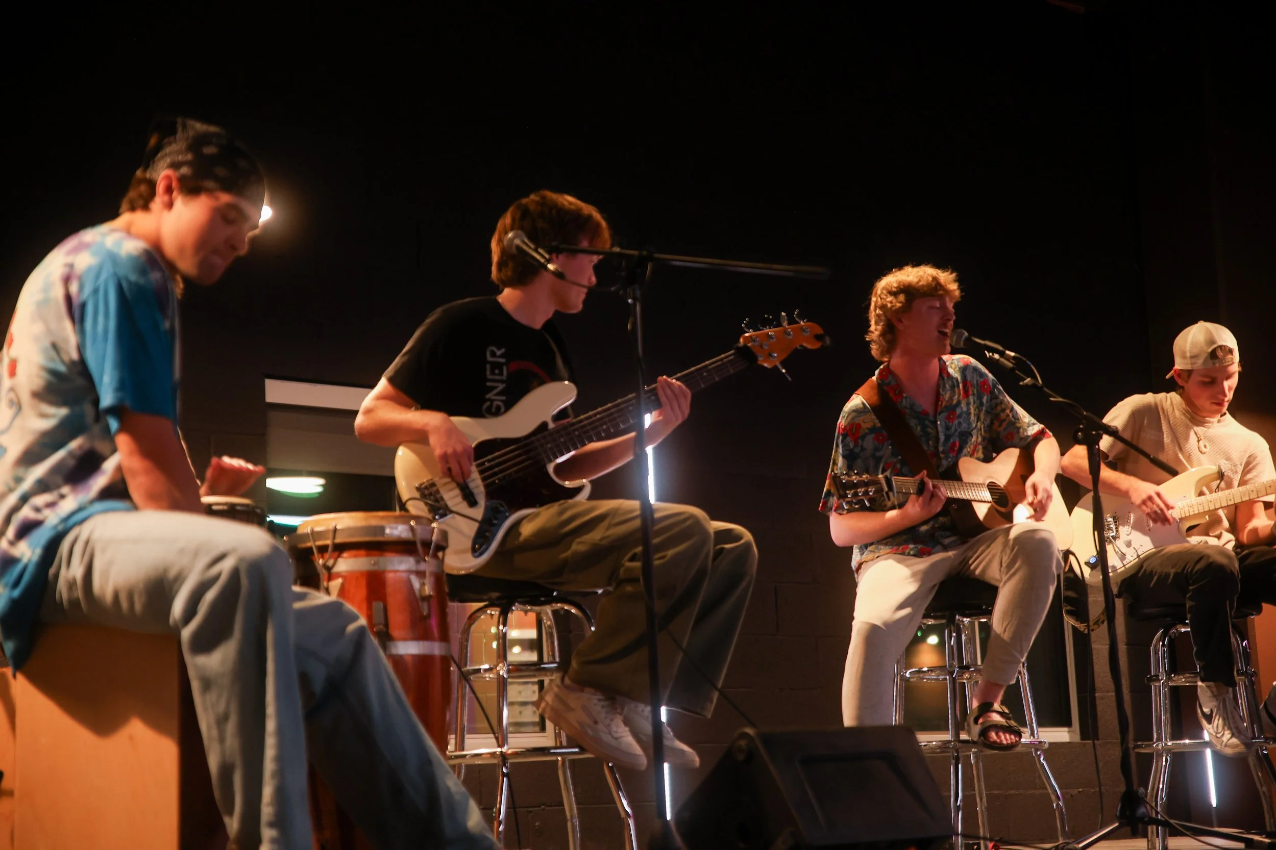 Four young men performing music on stage, playing drums, bass guitar, acoustic guitar, and electric guitar.