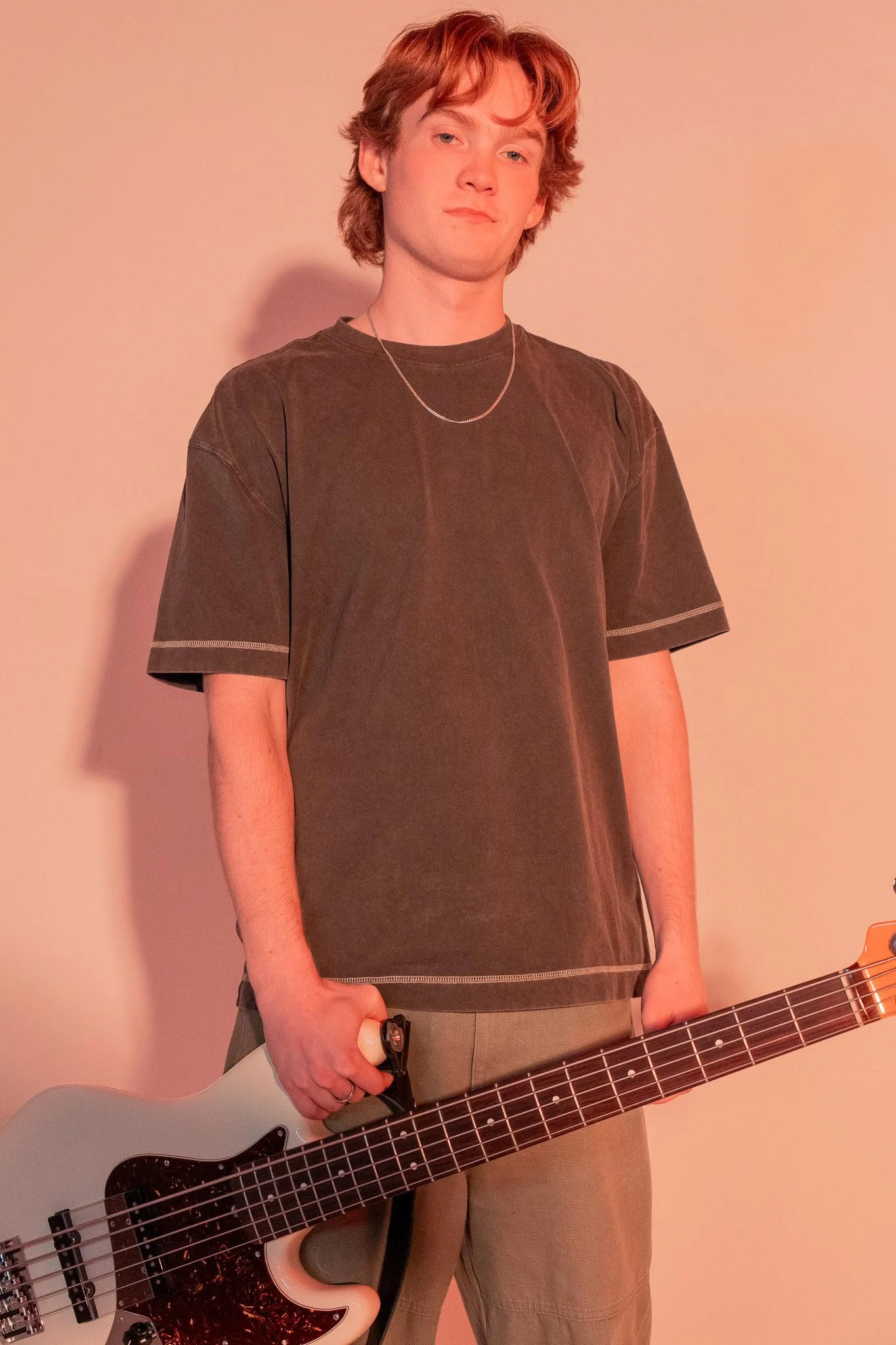 Young man with wavy red hair holding a white electric bass guitar, wearing a brown t-shirt and beige pants, standing against a pink background.