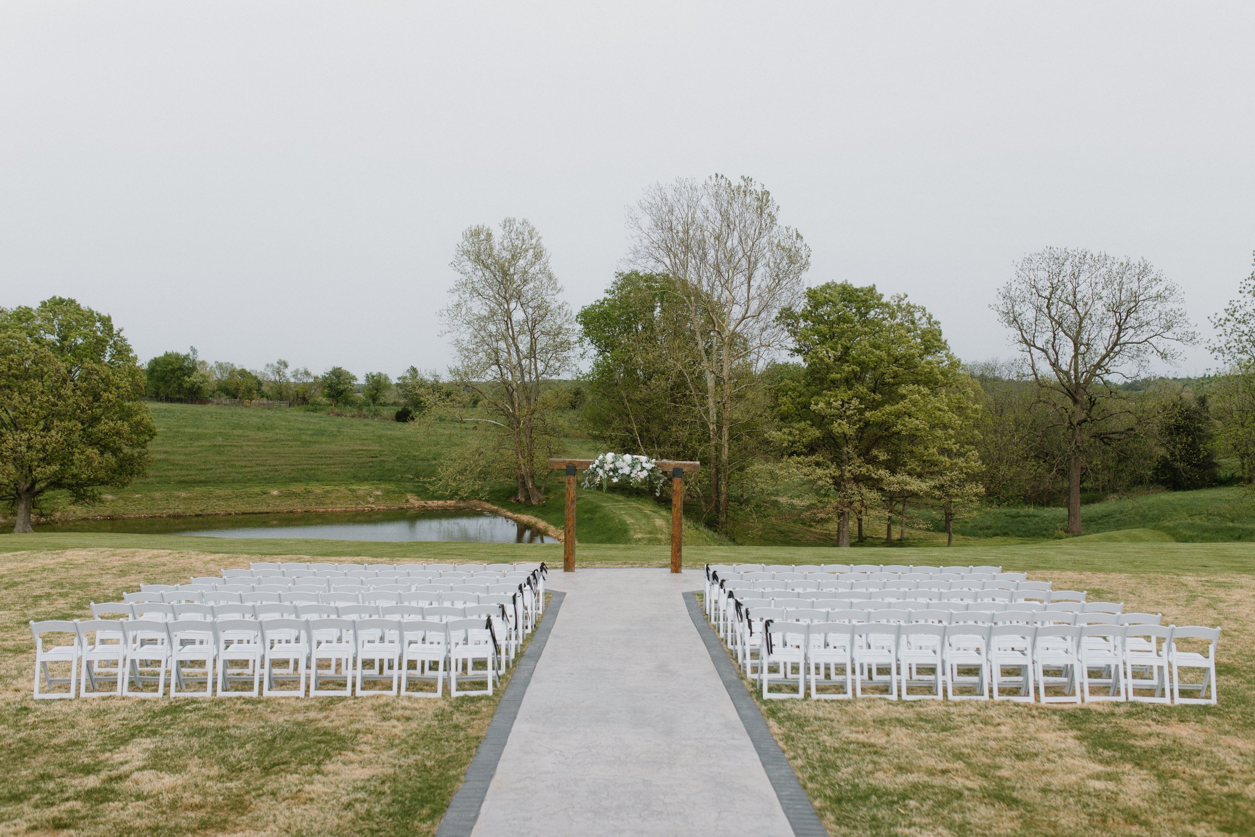 Outdoor wedding ceremony setup with white chairs arranged on either side of a concrete aisle, leading to a wooden arch decorated with white flowers, in a grassy field with trees and a small pond in the background.