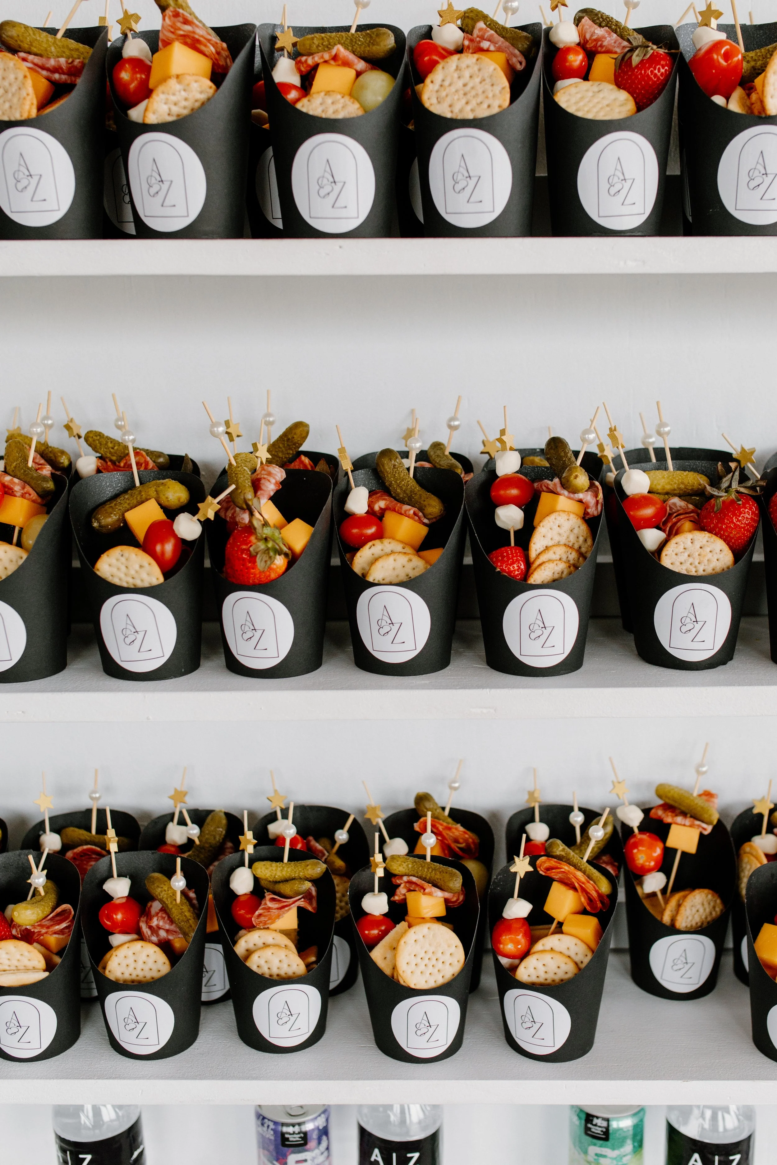 Three shelves filled with black paper cone containers holding cheese, cherry tomatoes, strawberries, crackers, pickles, and small skewers with star and pearl decorations, likely for a party or event.