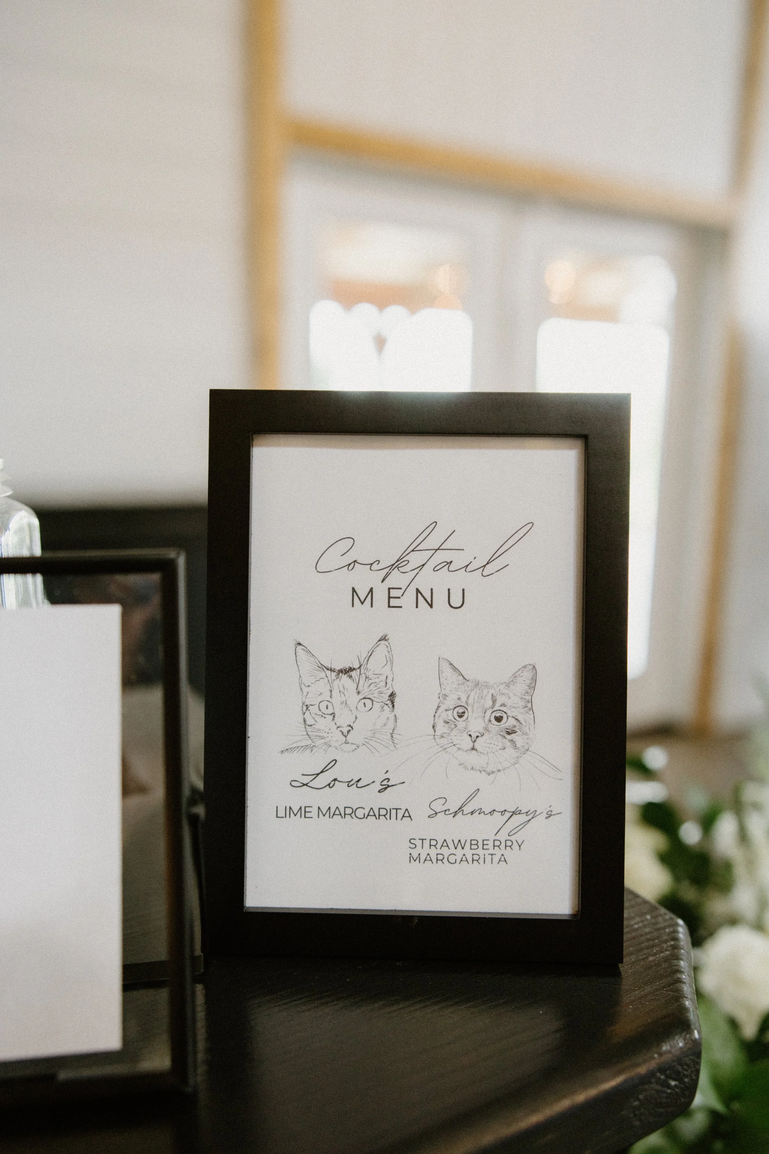 Framed cocktail menu featuring illustrations of two cats, listing drinks as Lime Margarita and Strawberry Margarita, placed on a dark wooden table.