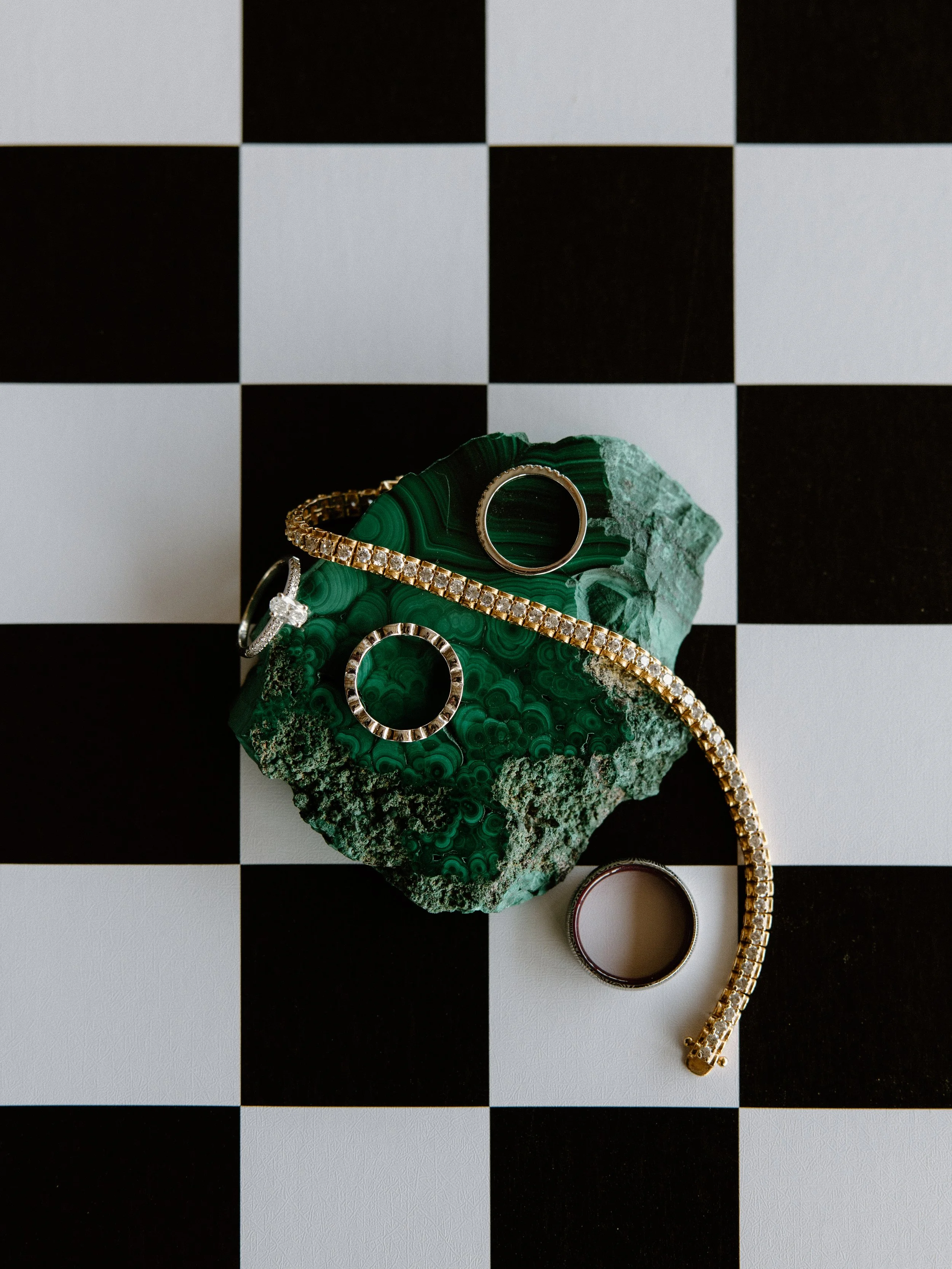 Jewelry including rings and a bracelet displayed on a green malachite stone on a black and white checkered surface.