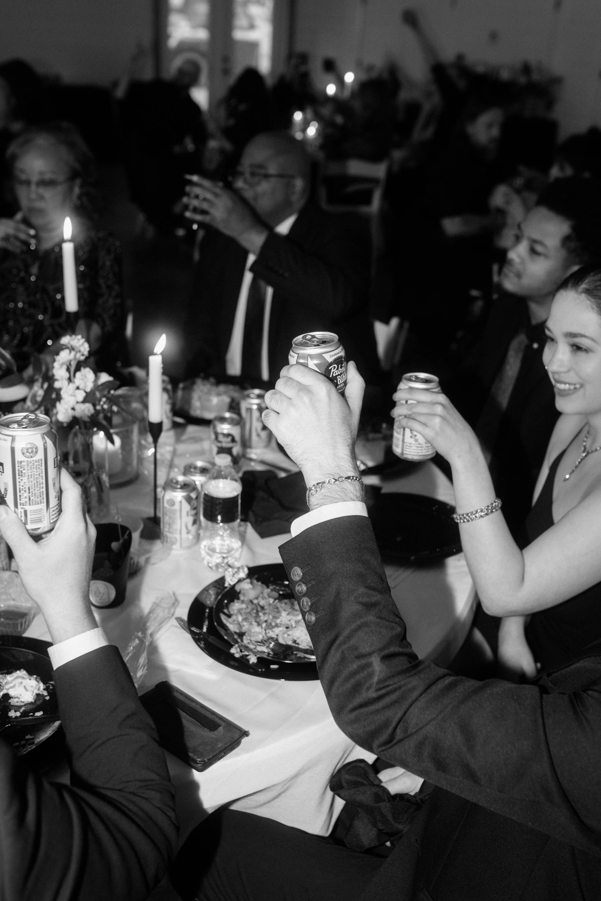 People toasting with cans of beer at a dinner party, with wine glasses and candles on the table.