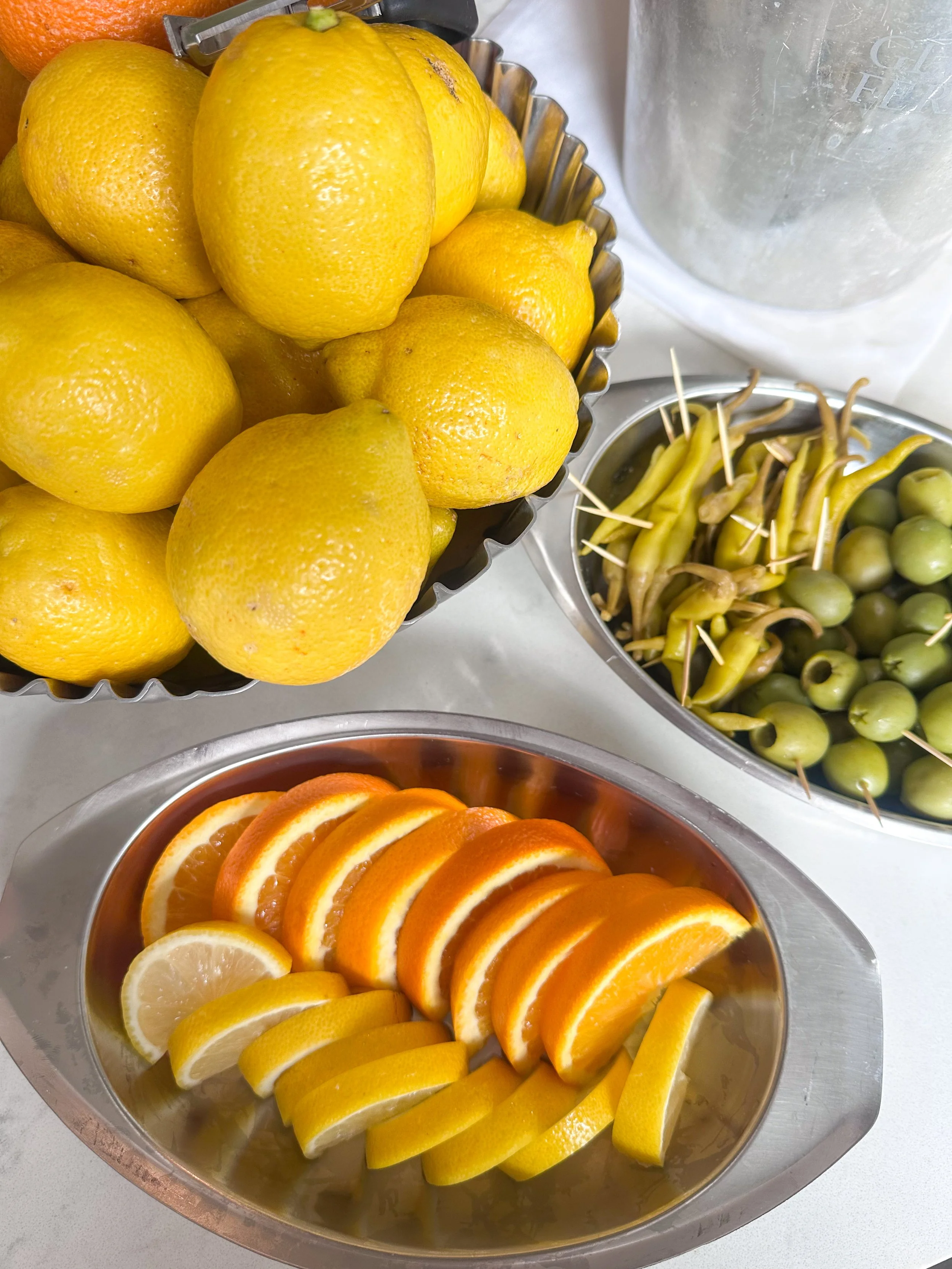 A bowl of yellow lemons, a bowl of green olives with toothpicks, and a tray of sliced oranges and lemons.