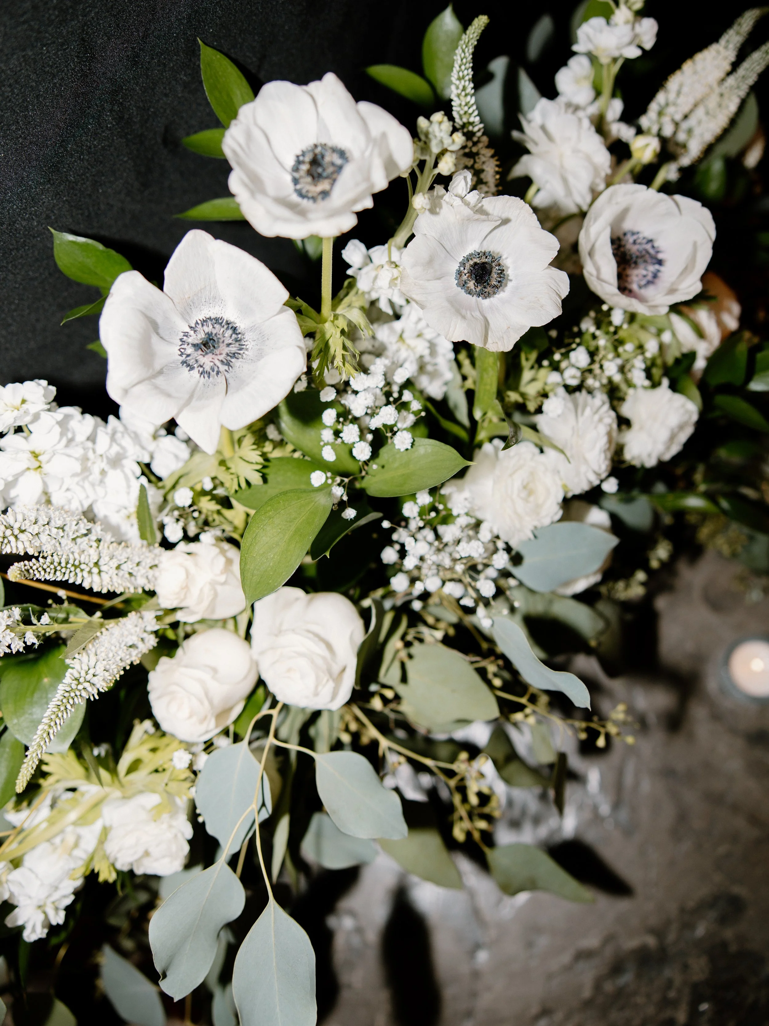 A bouquet of white flowers including poppies, roses, and various greenery, arranged against a dark background.