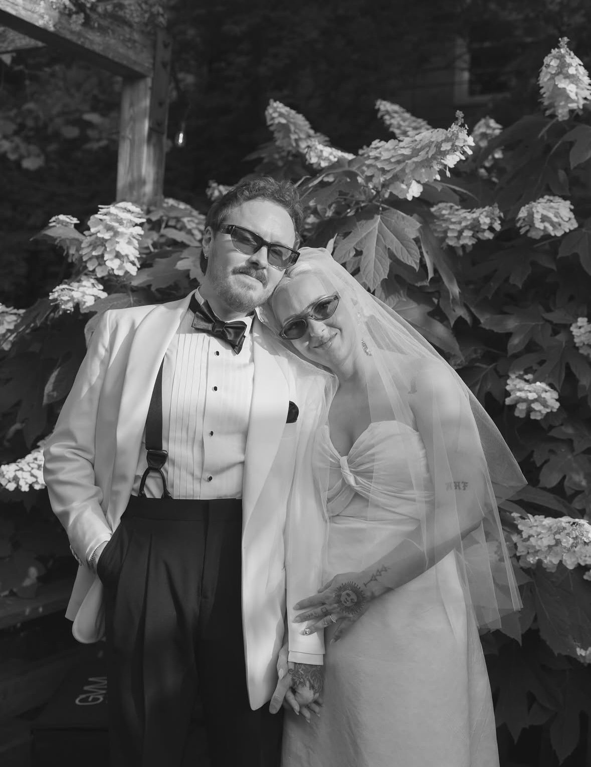 Black and white photo of a couple in wedding attire, standing close together outdoors with large leafy plants and flowers in the background, both wearing sunglasses.