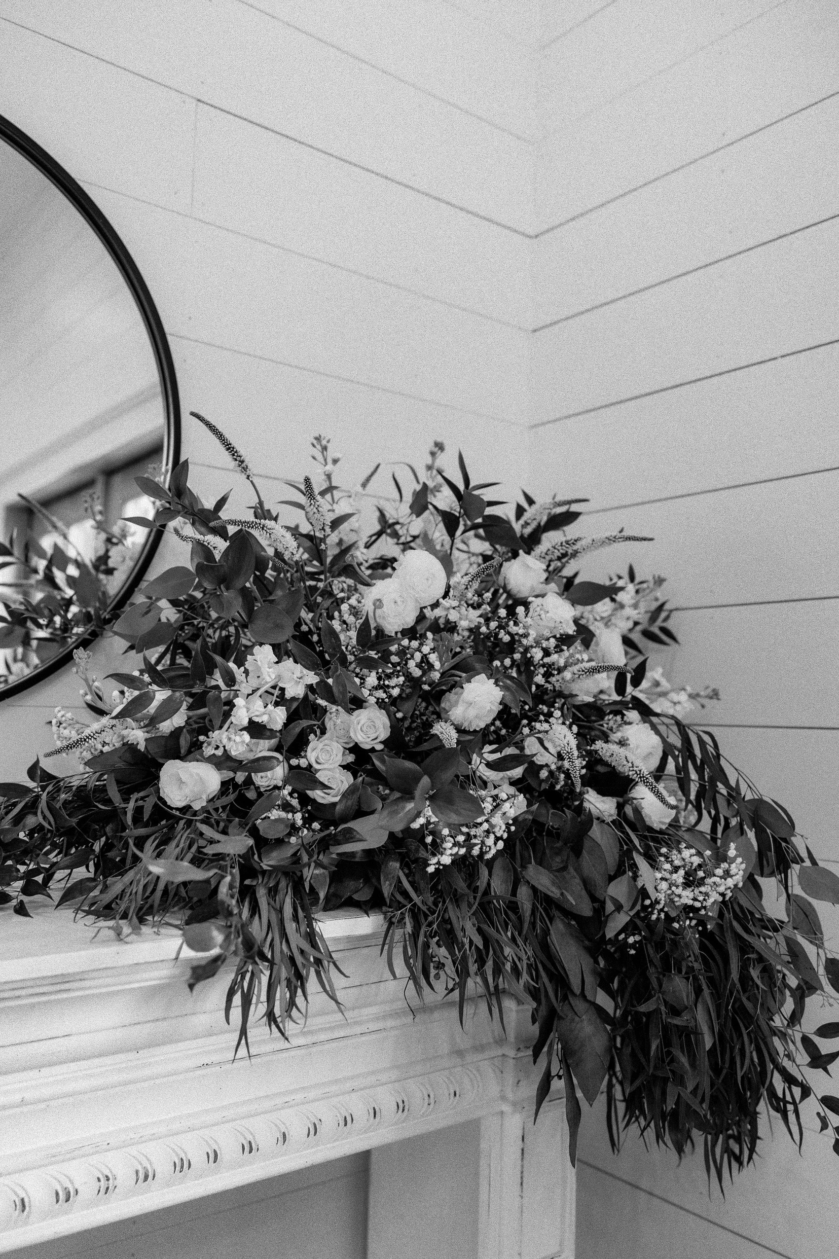 A black and white photo of a floral arrangement on a fireplace mantel, with a mirror reflecting part of the room in the background.