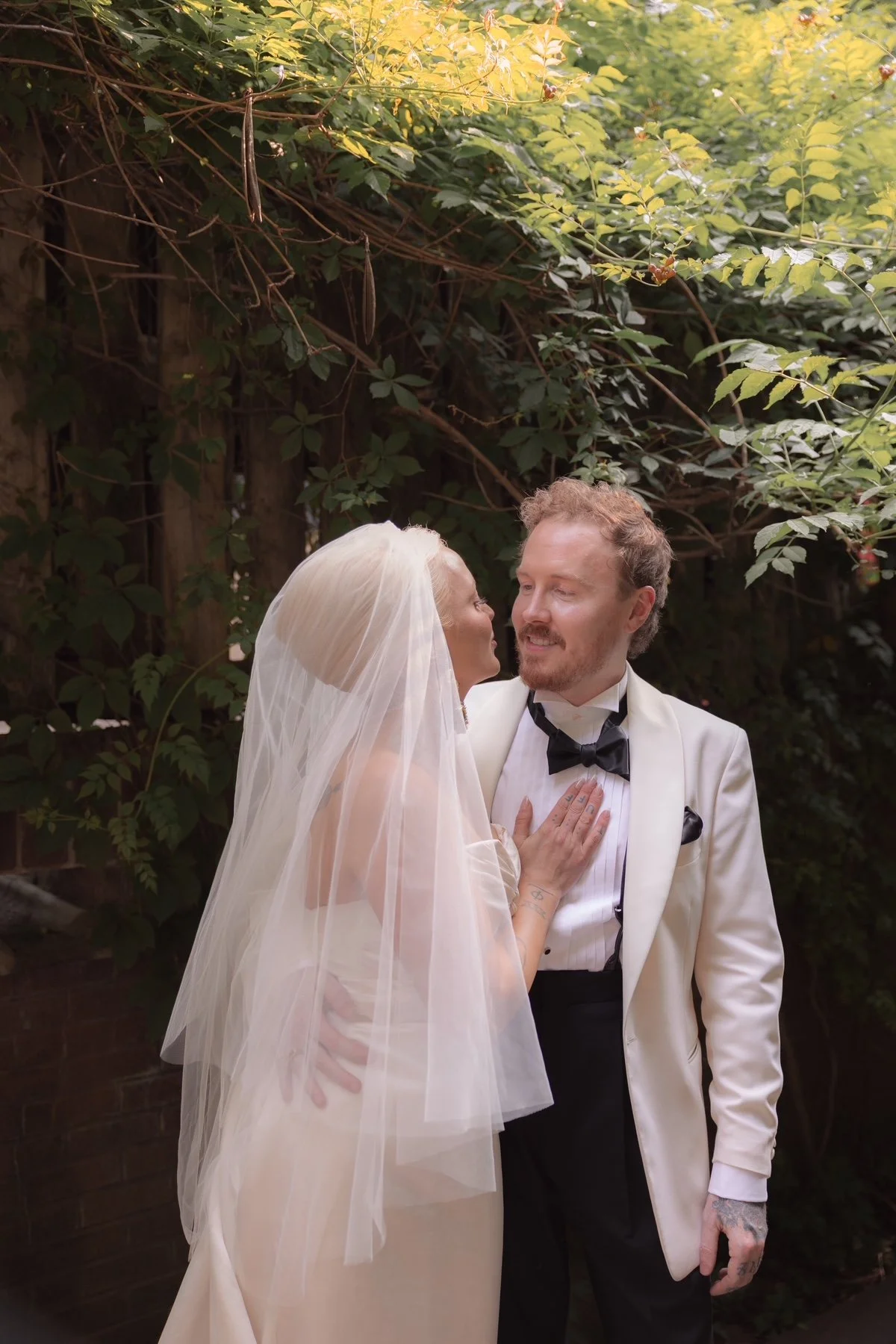 Bride and groom stand close together outdoors, the bride leaning towards the groom, who is looking at her affectionately. The bride is wearing a white wedding dress and veil, while the groom is dressed in a white tuxedo jacket with a black bow tie, in front of lush green foliage.