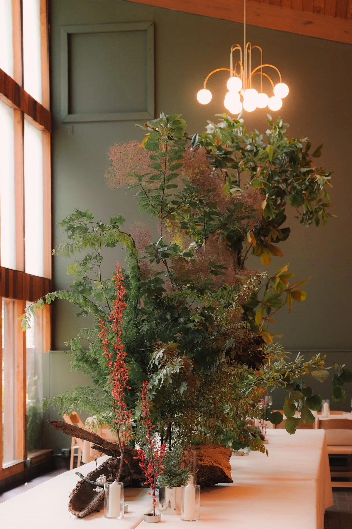 A large floral arrangement with green leaves, red flowers, and pink fluffy flowers sits on a white table in a room with green walls and wooden accents, illuminated by a modern chandelier with spherical light bulbs.
