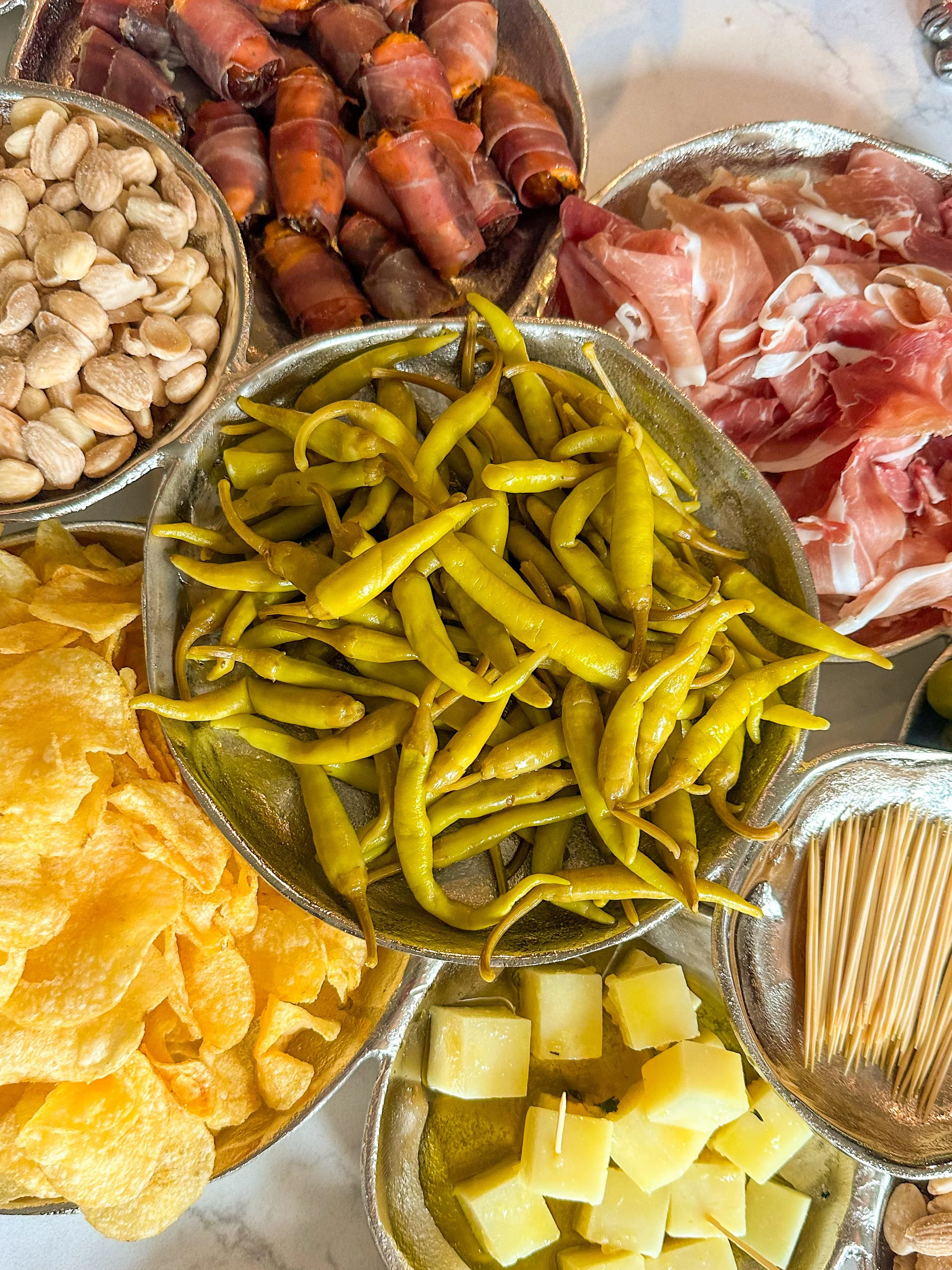 Assorted appetizer ingredients including green chili peppers, cheese cubes, potato chips, cured meats, green beans, nuts, and breadsticks displayed in metal bowls.