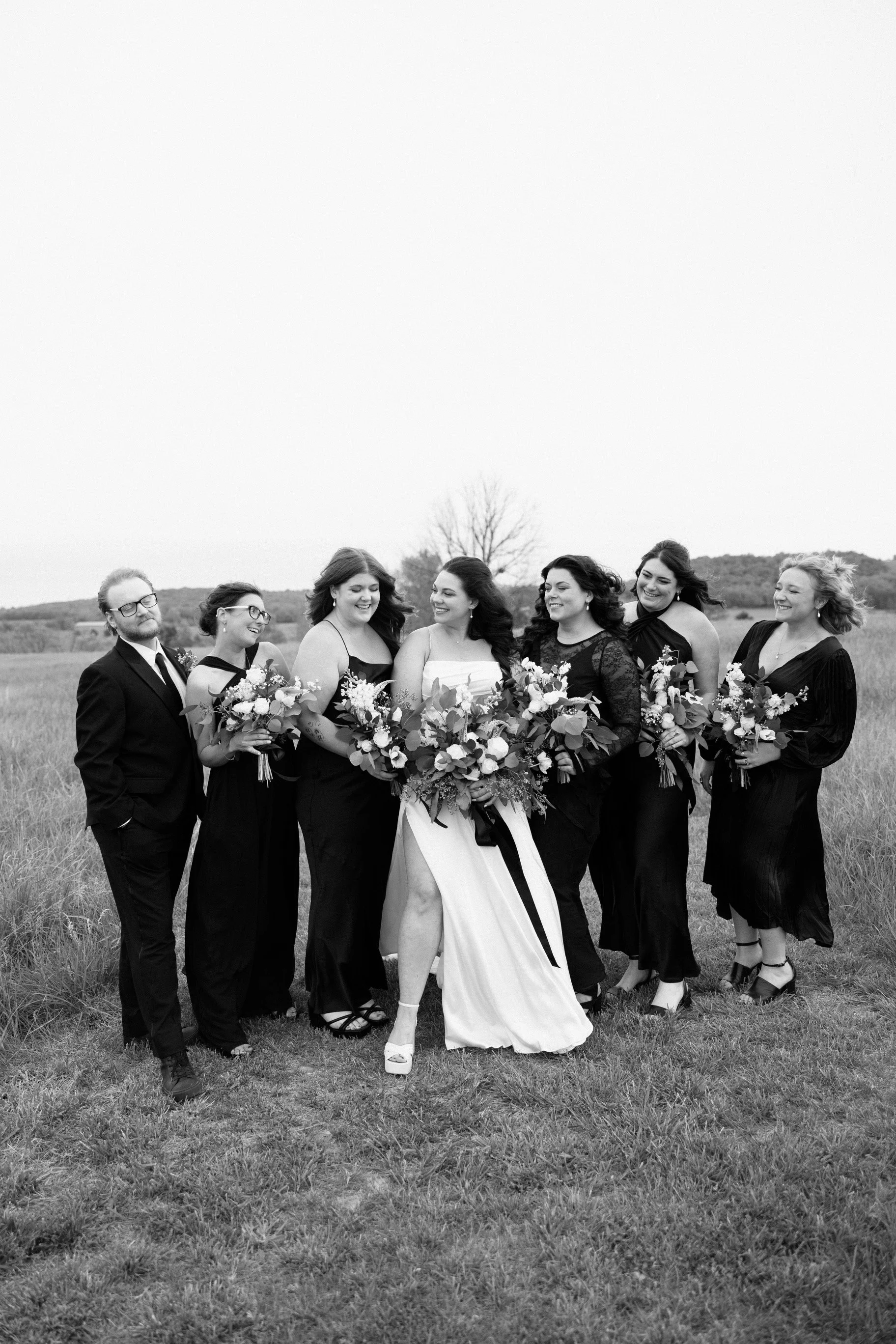 Black and white photo of a bride and six bridesmaids in a grassy field, holding bouquets and smiling.