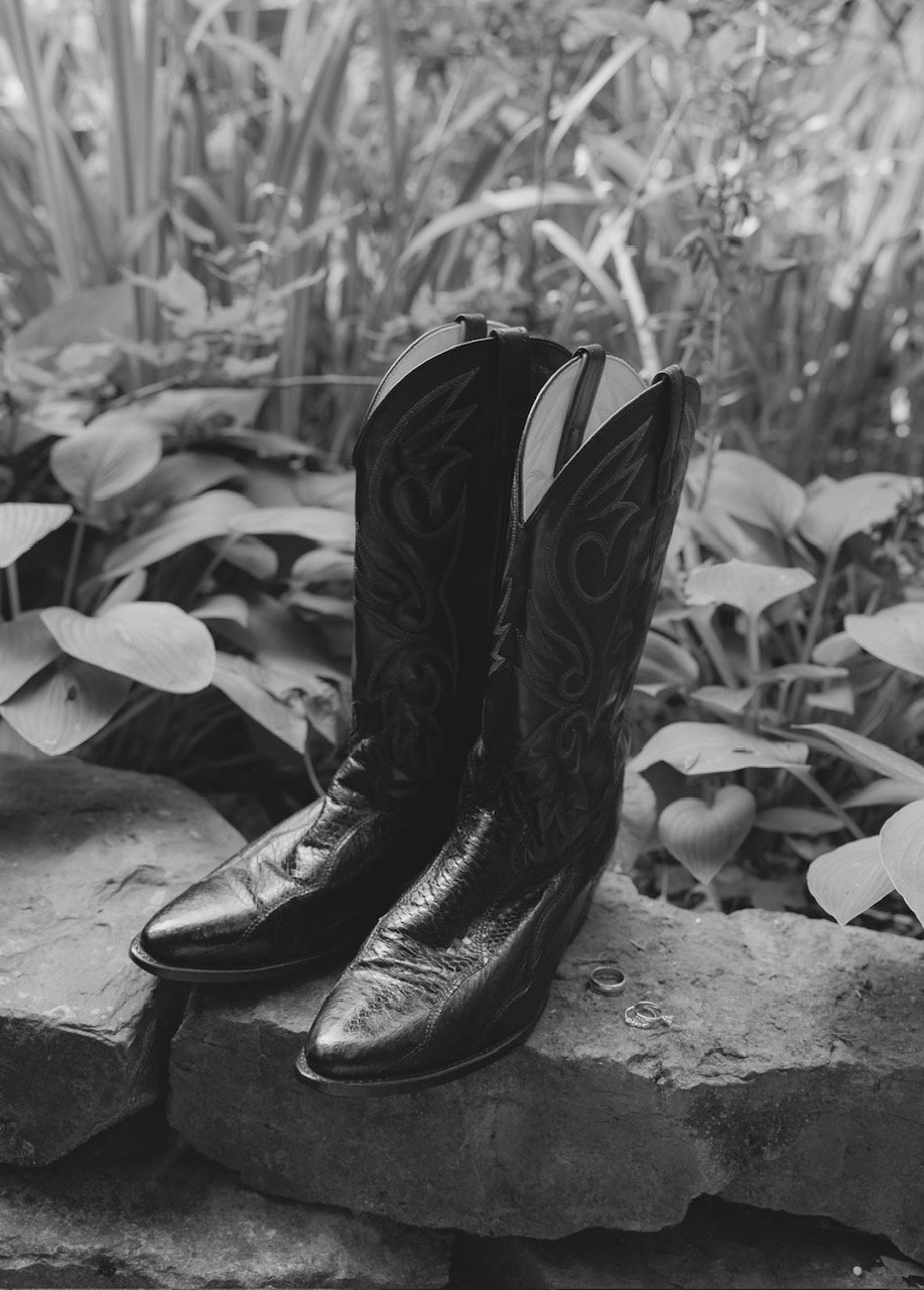 Black cowboy boots with embroidered designs placed on a stone ledge outdoors with plants in the background.