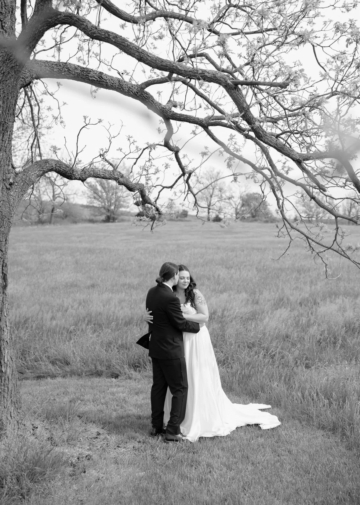 Black and white photo of a bride and groom embracing under a large tree in a open field.