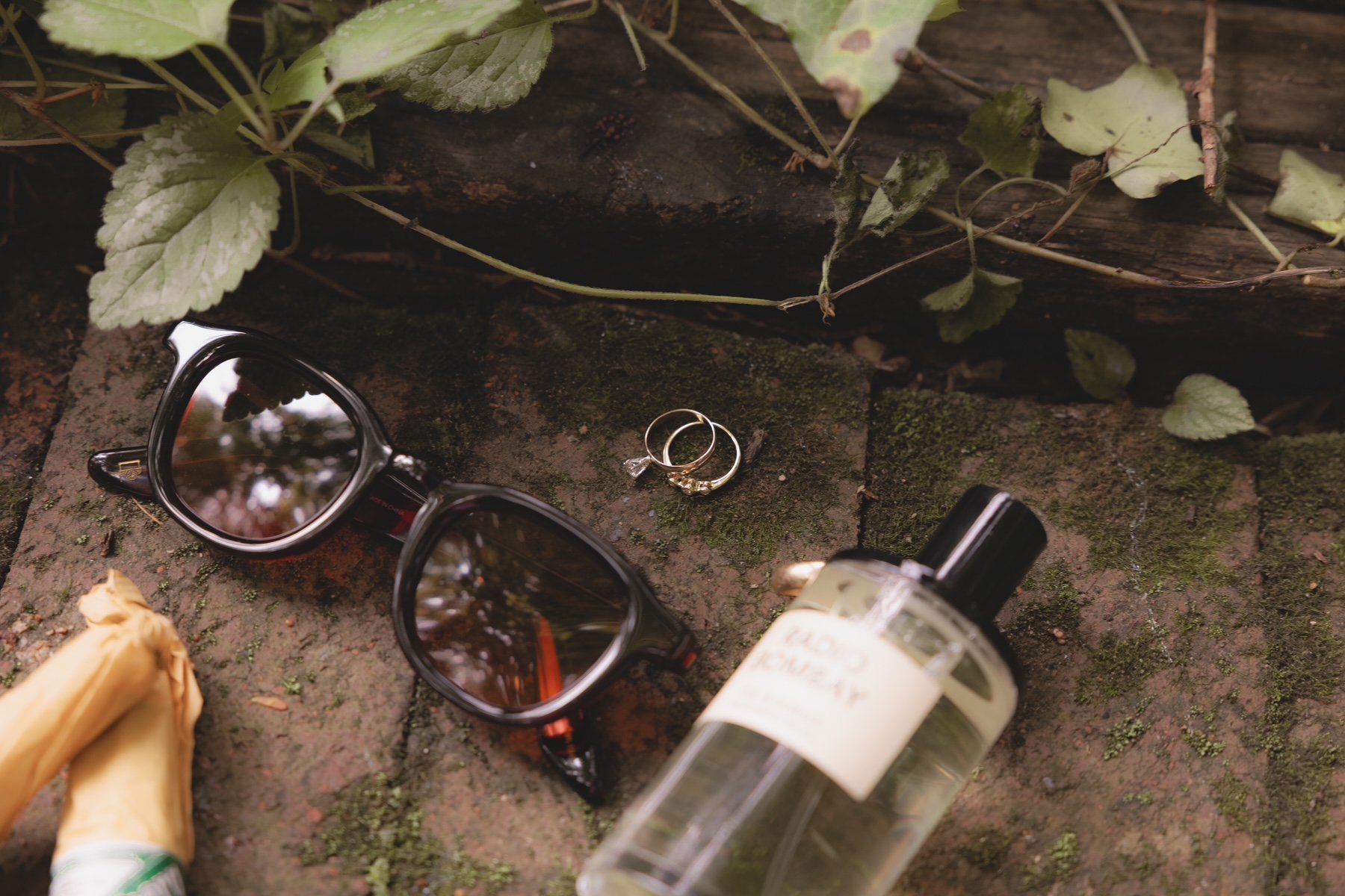 Sunglasses, two rings, a perfume bottle, and a yellow flower on a mossy brick surface outdoors.