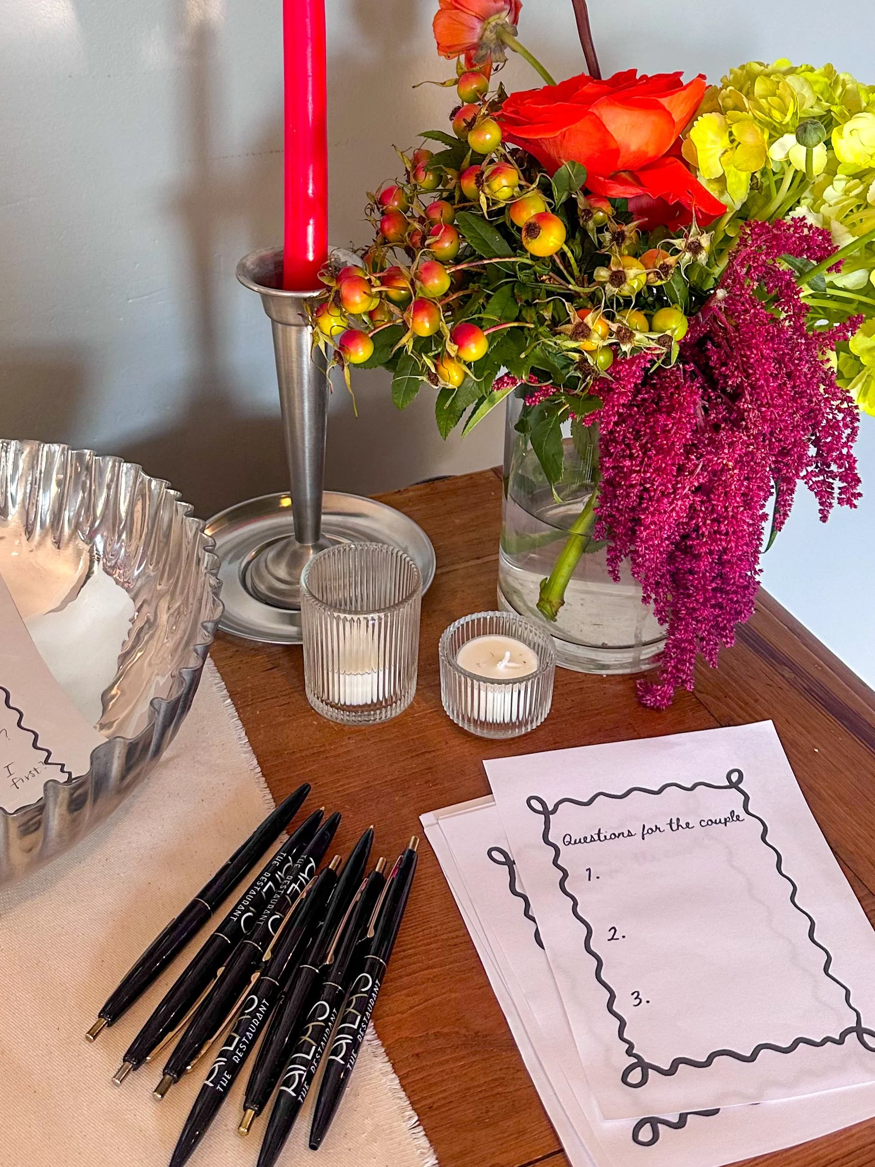 Decorative table arrangement with a tall red candle in a silver candlestick, a glass vase filled with colorful flowers and berry branches, two small candles in glass holders, a stack of papers titled 'Questions for the couple', and several black pens.