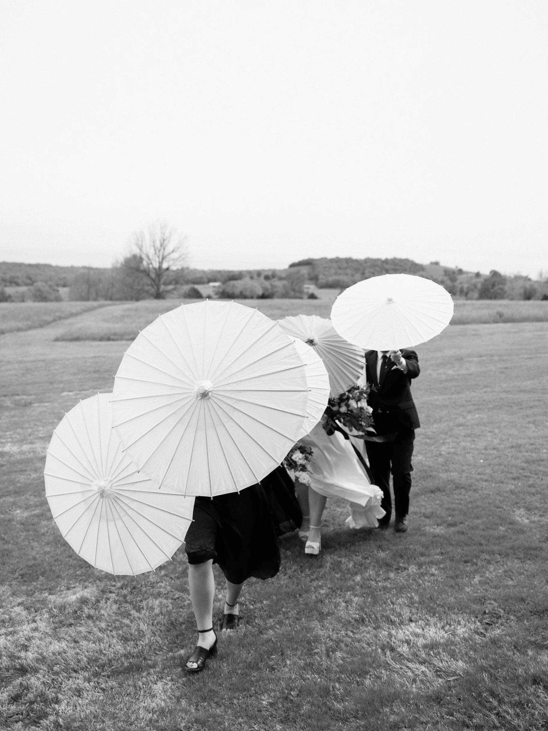 Black and white photo of a wedding party walking across a grassy field, with people holding parasols to shield from the sun. The bride is dressed in a wedding gown and carrying a bouquet, with one person in a tuxedo beside her.