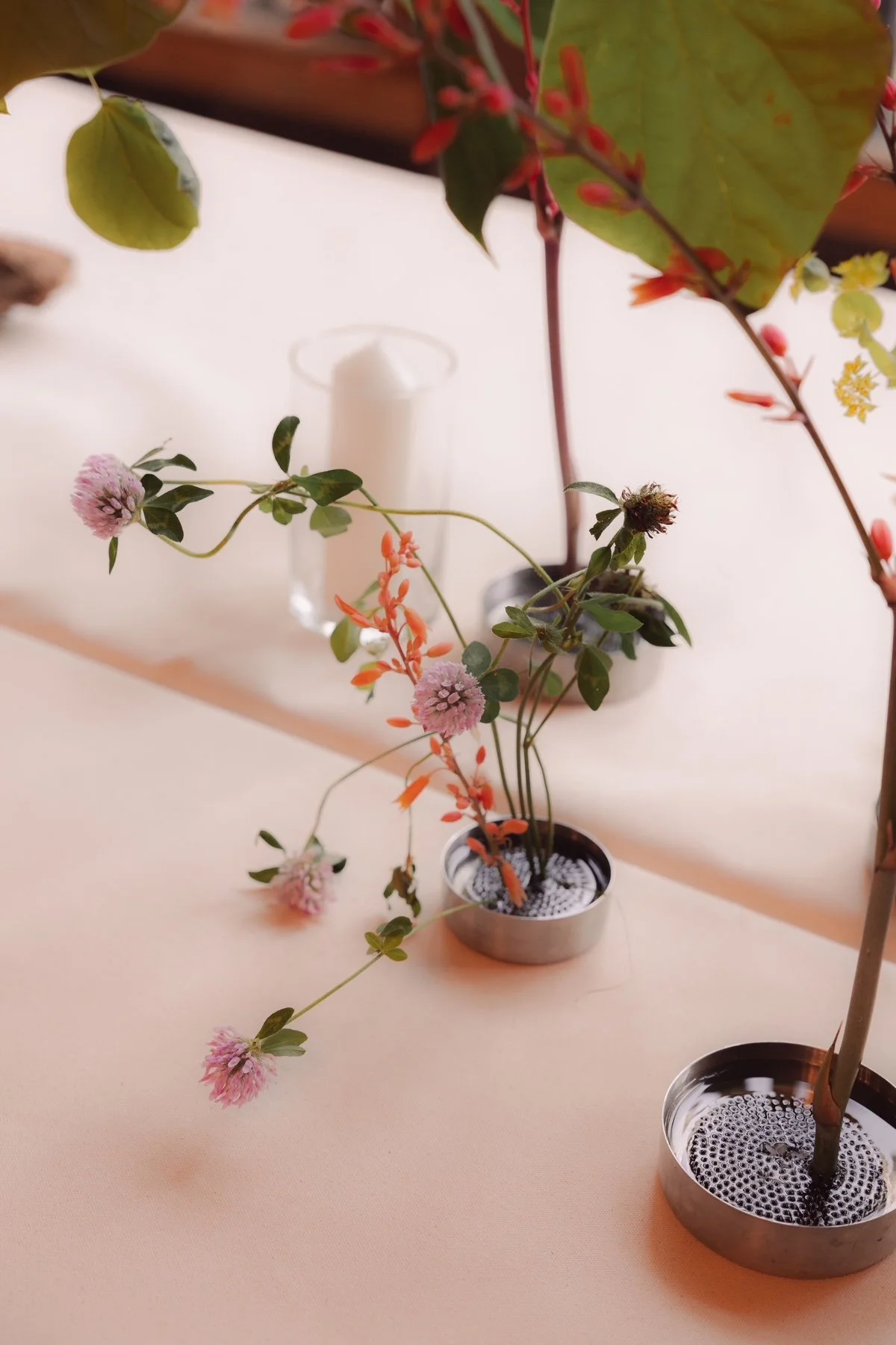 Vase with pink and purple flowers on a light pink table, with a candle and other plants in the background.