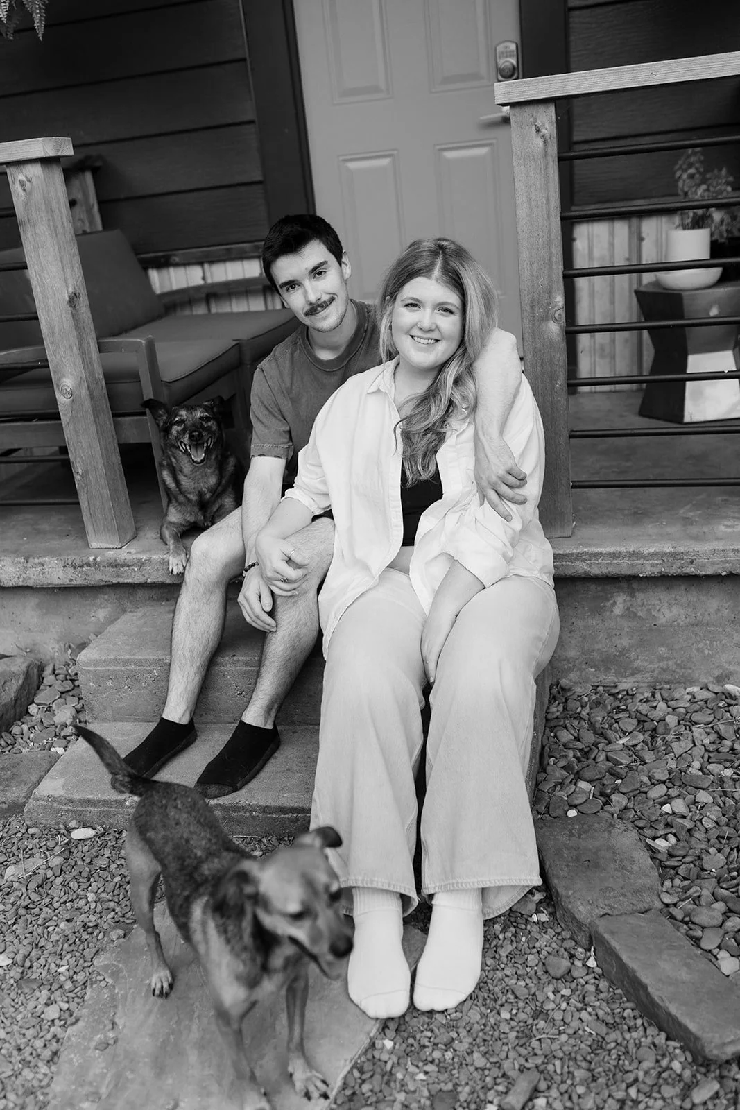 A smiling woman sitting on porch steps, with a young man sitting beside her, and two dogs - one sitting behind them and another standing on gravel in front. The porch has a wooden railing, a door, and potted plants.