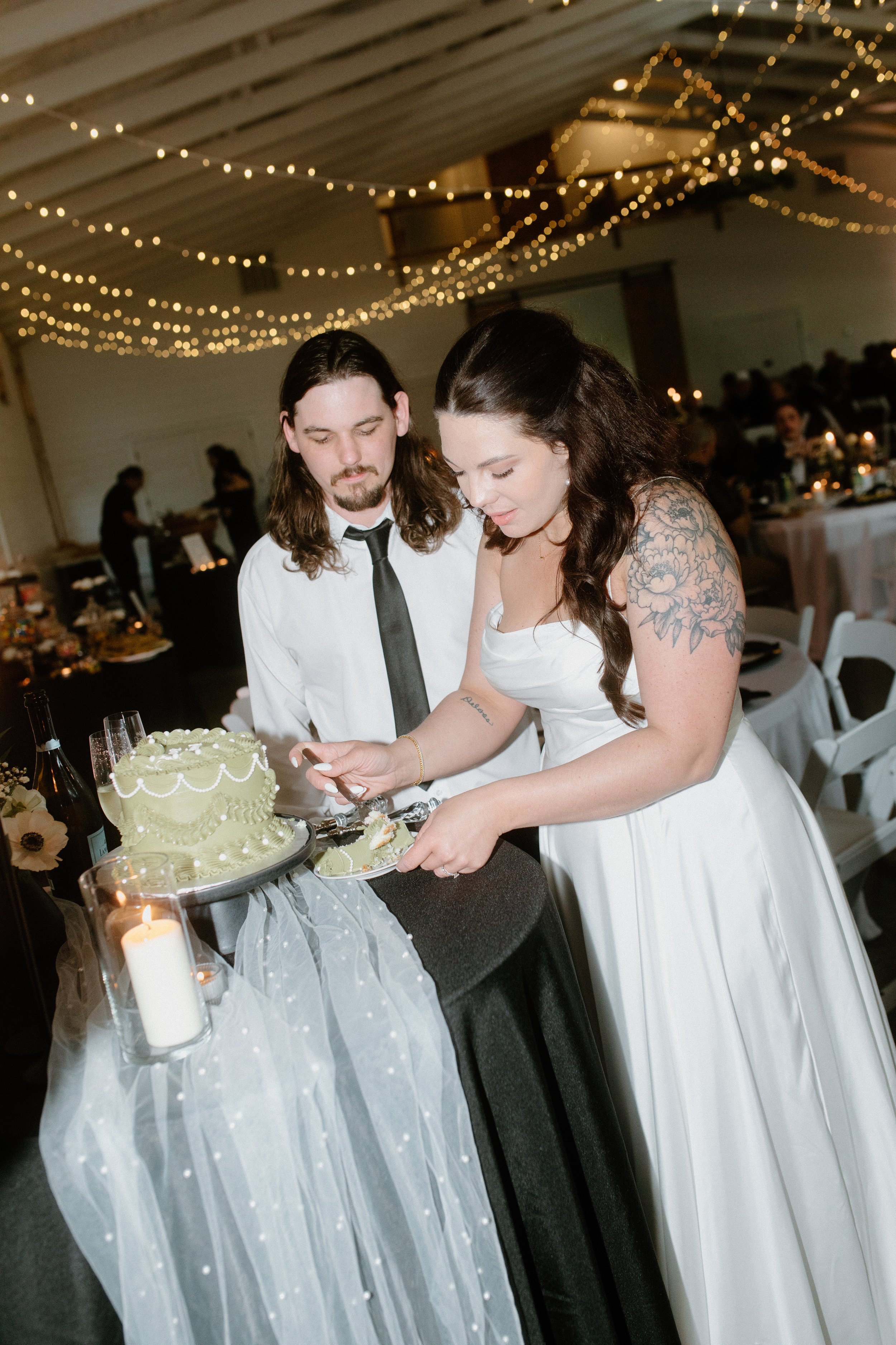A bride and groom cutting a wedding cake together at their reception, with fairy lights hanging from the ceiling and tables decorated with candles and floral arrangements in the background.