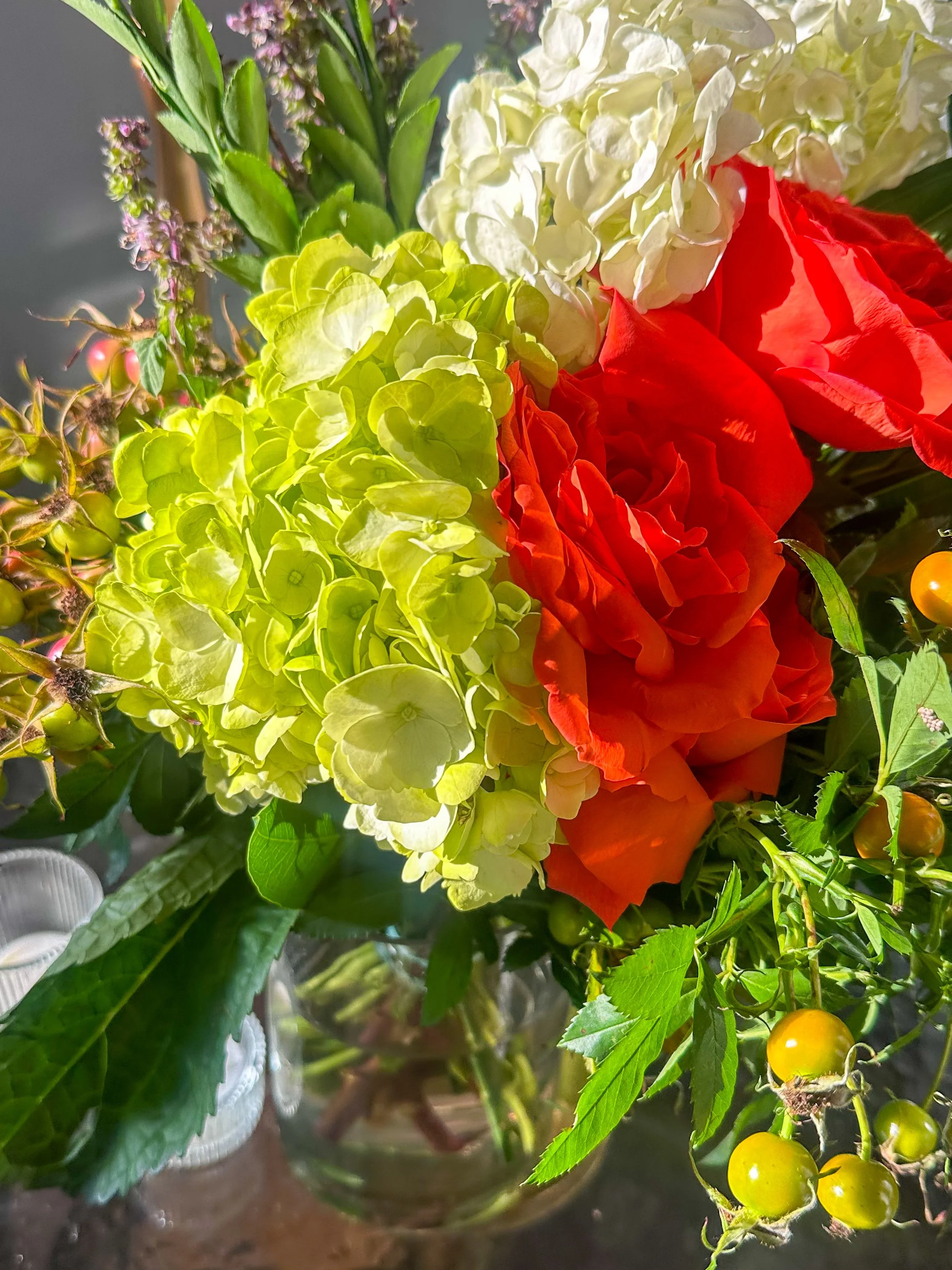 Close-up of a colorful flower arrangement in a glass vase, featuring lime green hydrangeas, bright orange roses, white hydrangeas, with some purple and green foliage and small yellow berries.