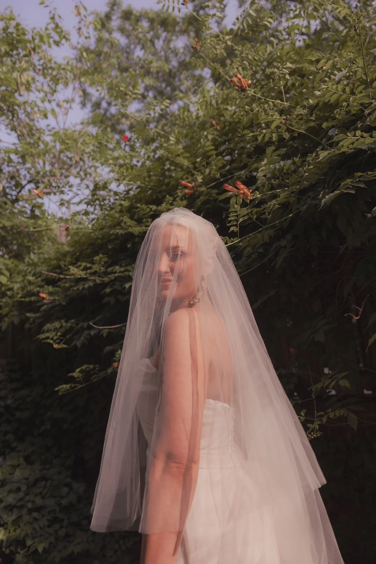 A woman in a wedding dress with a veil standing outdoors amidst green foliage and pink flowers.