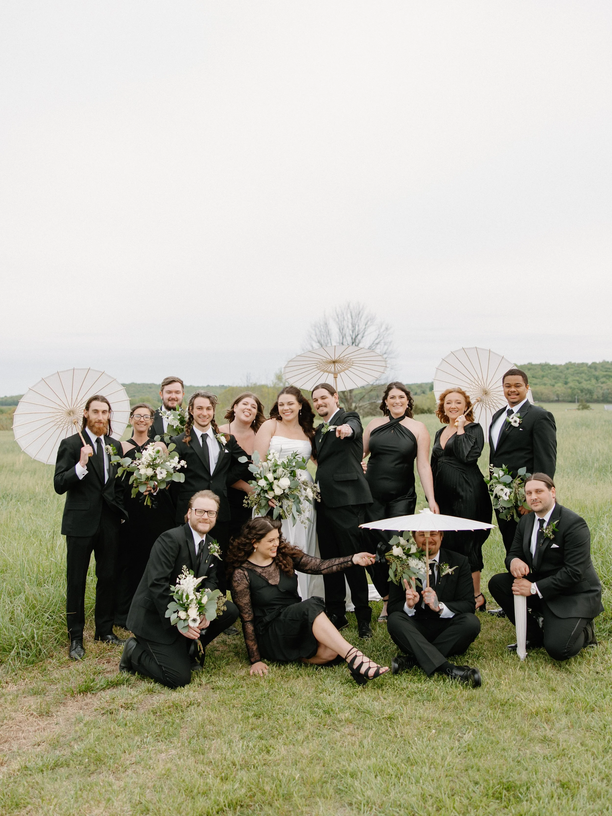 A large wedding party standing and sitting in an open grassy field with a cloudy sky, holding umbrellas and bouquets, dressed in formal black and white attire.