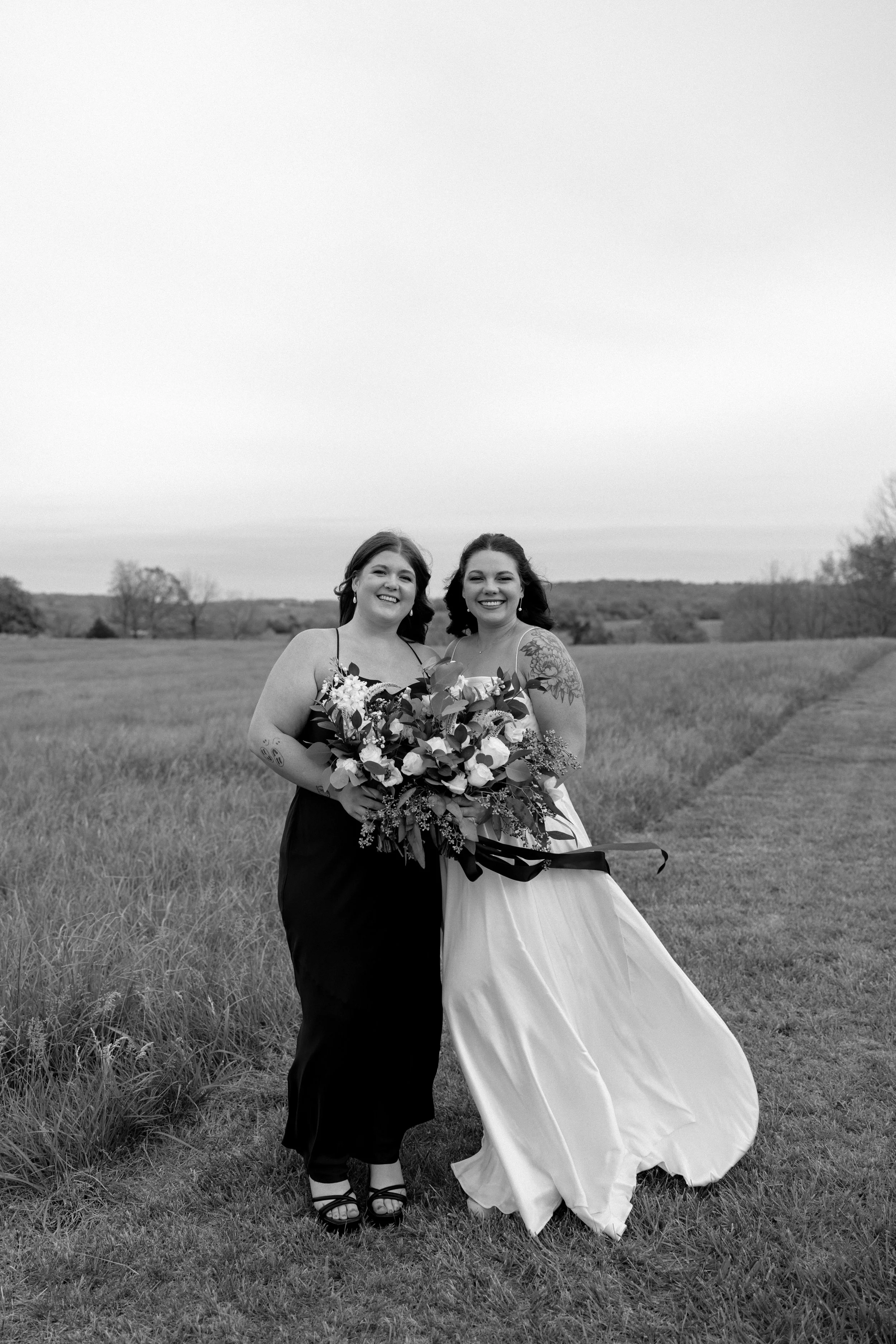 Two women standing in a field, smiling, one in a black dress holding a large bouquet of flowers, the other in a white wedding dress.