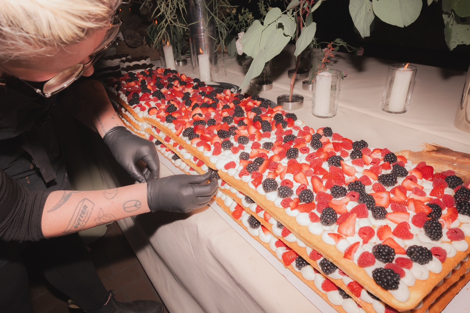 Person wearing black gloves decorating a long dessert with strawberries, blackberries, and cream, on a table with candles and greenery.
