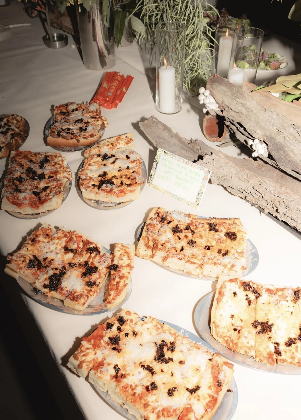 Various slices of homemade margherita pizza on plates arranged on a white table at a buffet or event setup, with candles, greenery, and bark decorations in the background.