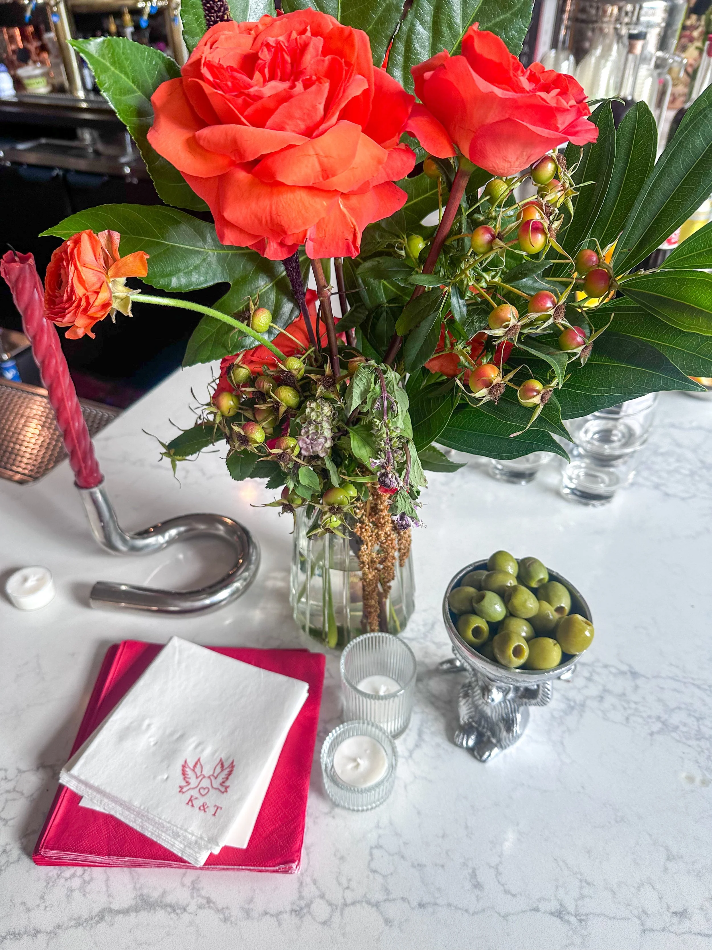 A floral arrangement with red-orange roses, green berries, and large green leaves in a glass vase. On the table, there are pink and white napkins with a logo, two small candles, and a bowl of green olives. There is also a pink candle in a silver holder and a marble countertop.