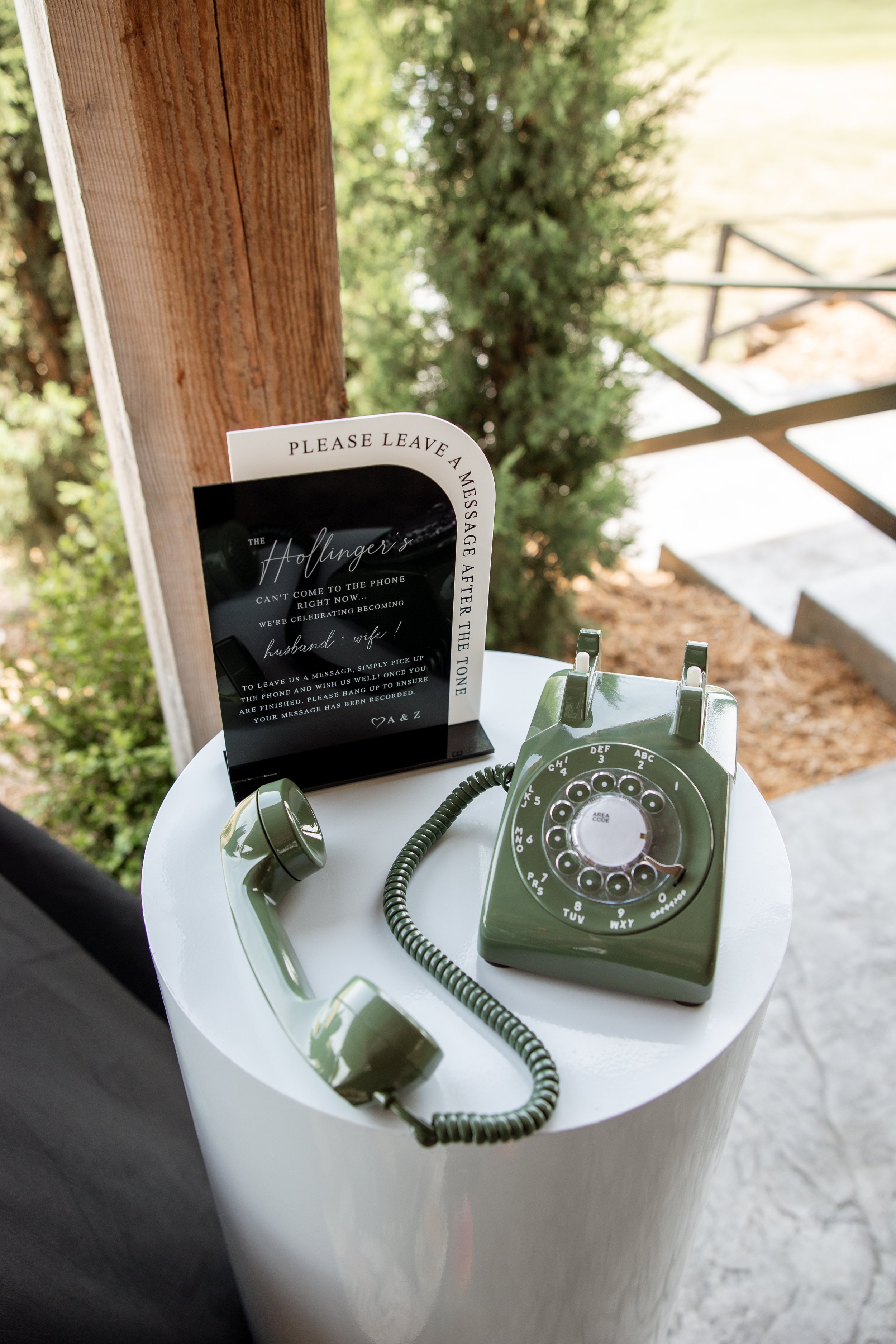 A vintage green rotary telephone on a white round table, with a sign requesting guests to leave a message after the tone at a wedding or event.