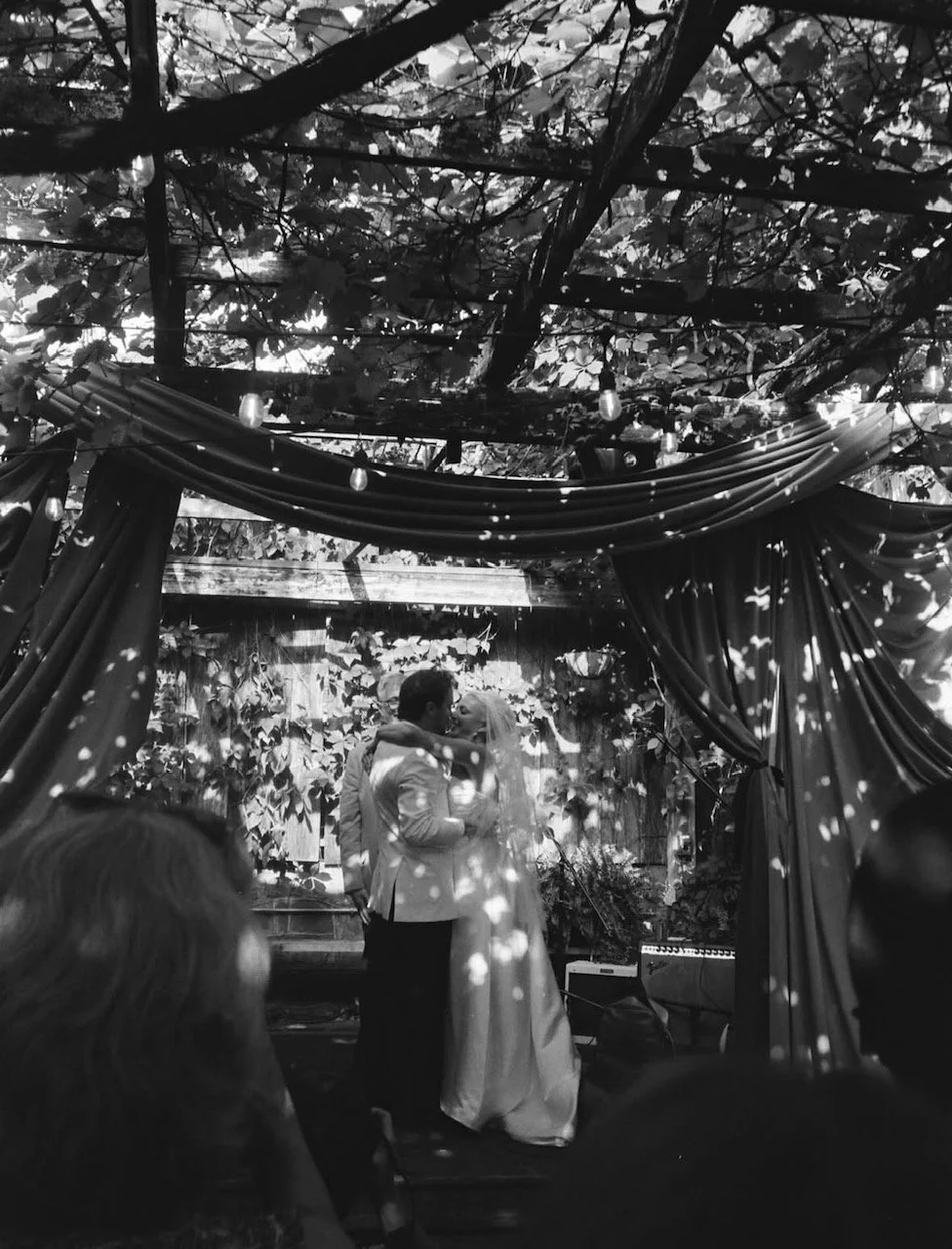 A black-and-white photo of a bride and groom sharing a kiss during their wedding ceremony. They are standing in a decorated outdoor venue with draped curtains and string lights, surrounded by guests.