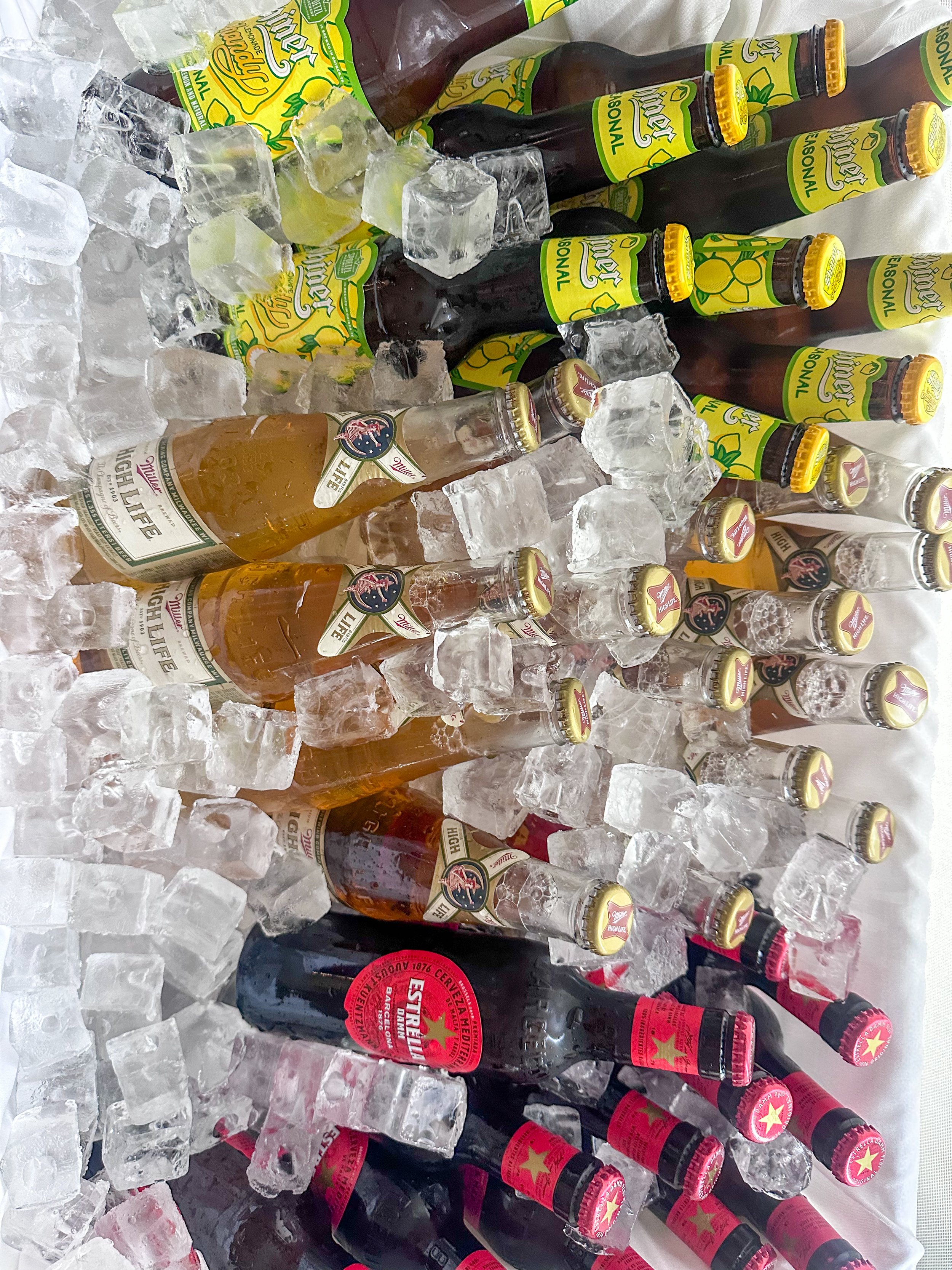 Several bottles of beer displayed on ice, including brands with green and yellow labels, and darker bottles with red labels. The bottles are on a white surface with ice cubes scattered around.