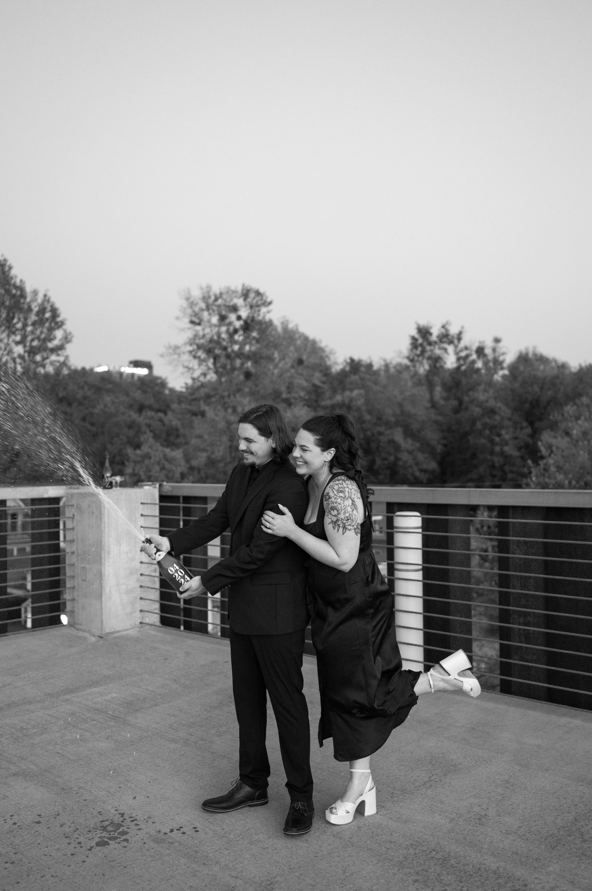 A man and a woman in formal attire celebrating on a rooftop with trees in the background, opening a champagne bottle with champagne spraying out.