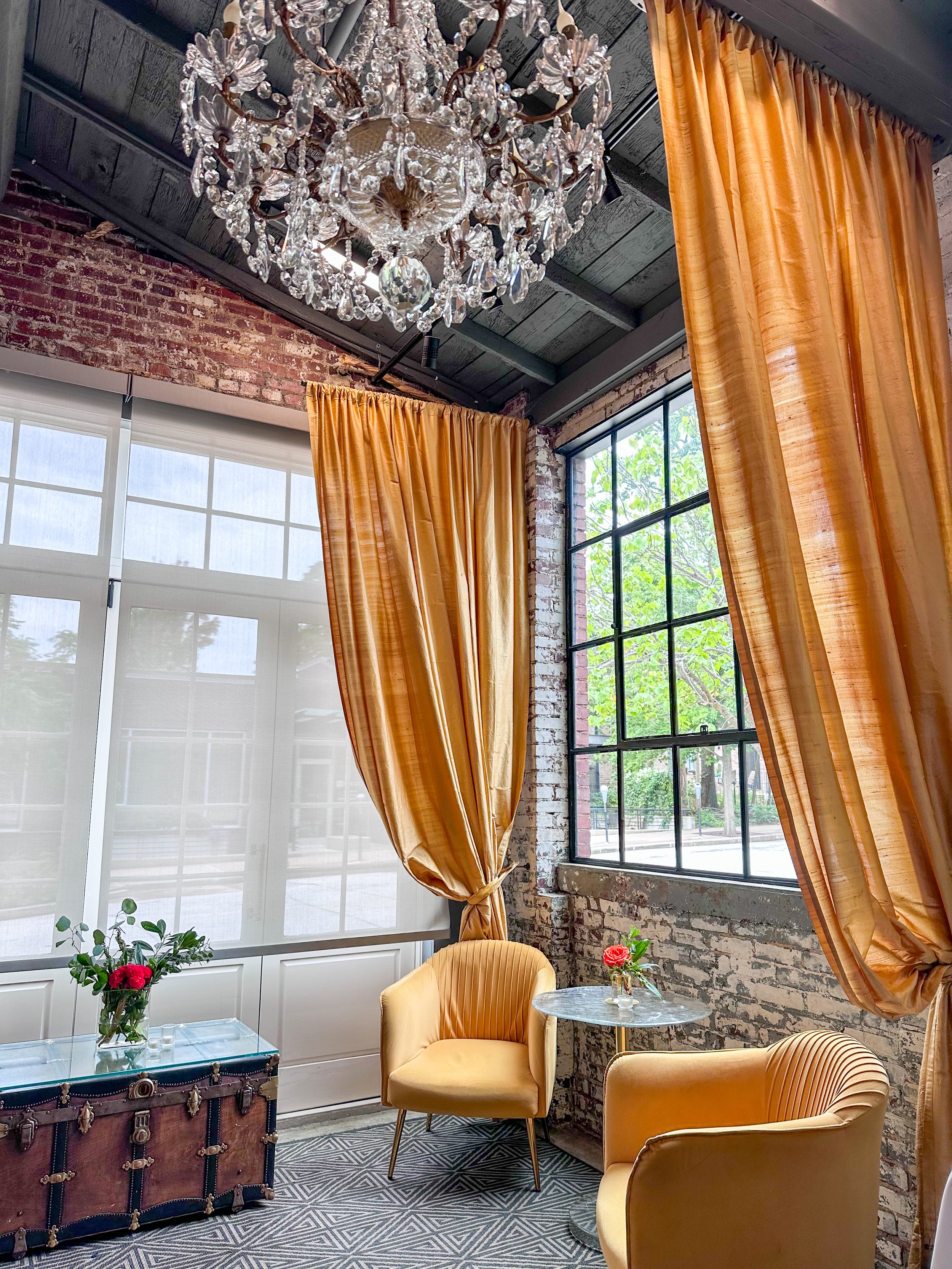 A cozy interior space with a crystal chandelier, large windows with black frames, golden curtains, two yellow armchairs, a small round table with a flower vase, a vintage trunk with a glass top and green plants, brick walls, and patterned carpet.