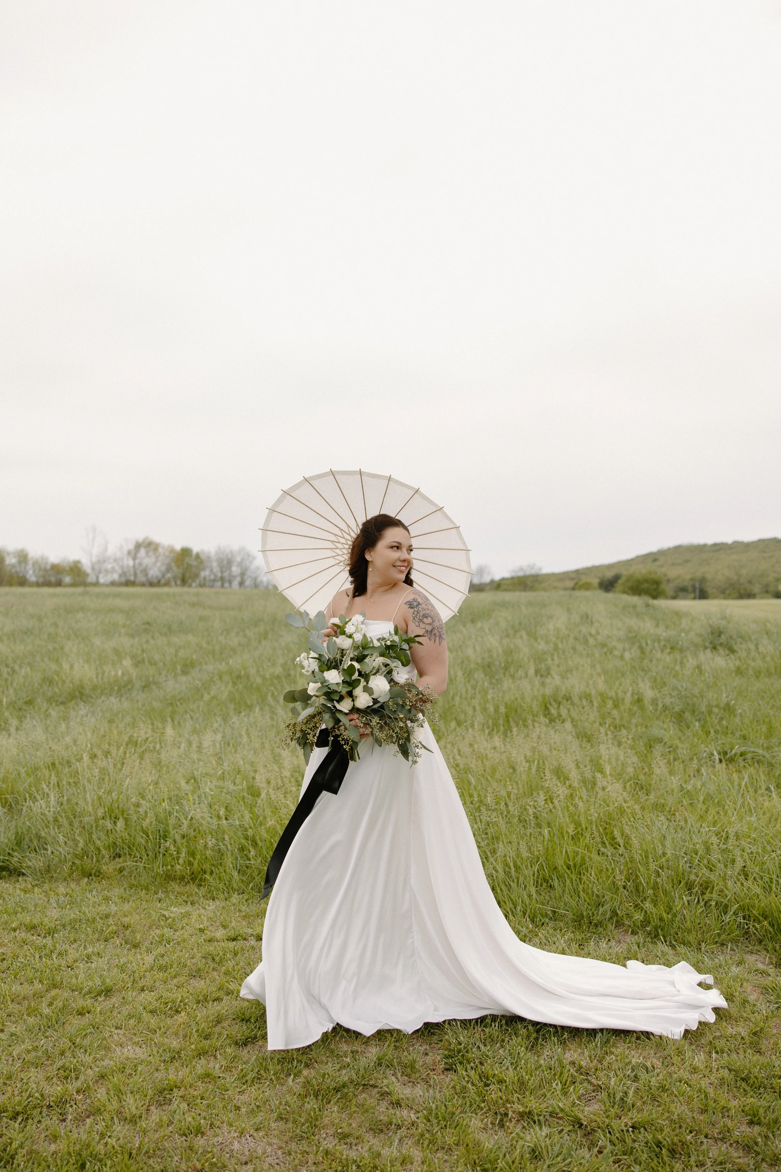 A woman in a white wedding dress holding a large bouquet, standing in a green field with an umbrella behind her, and smiling while looking to the side.