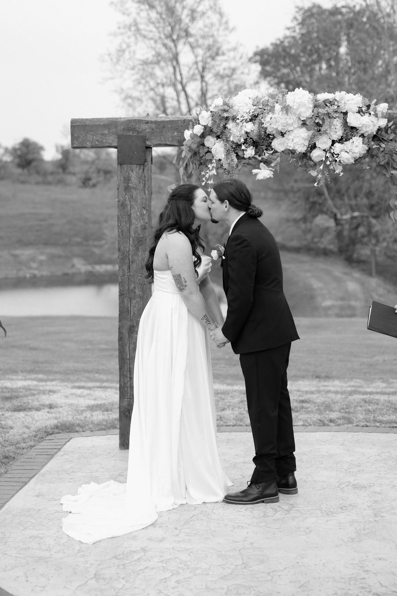 A wedding ceremony with a couple kissing under a floral arch, outdoors with trees in the background, in black and white.