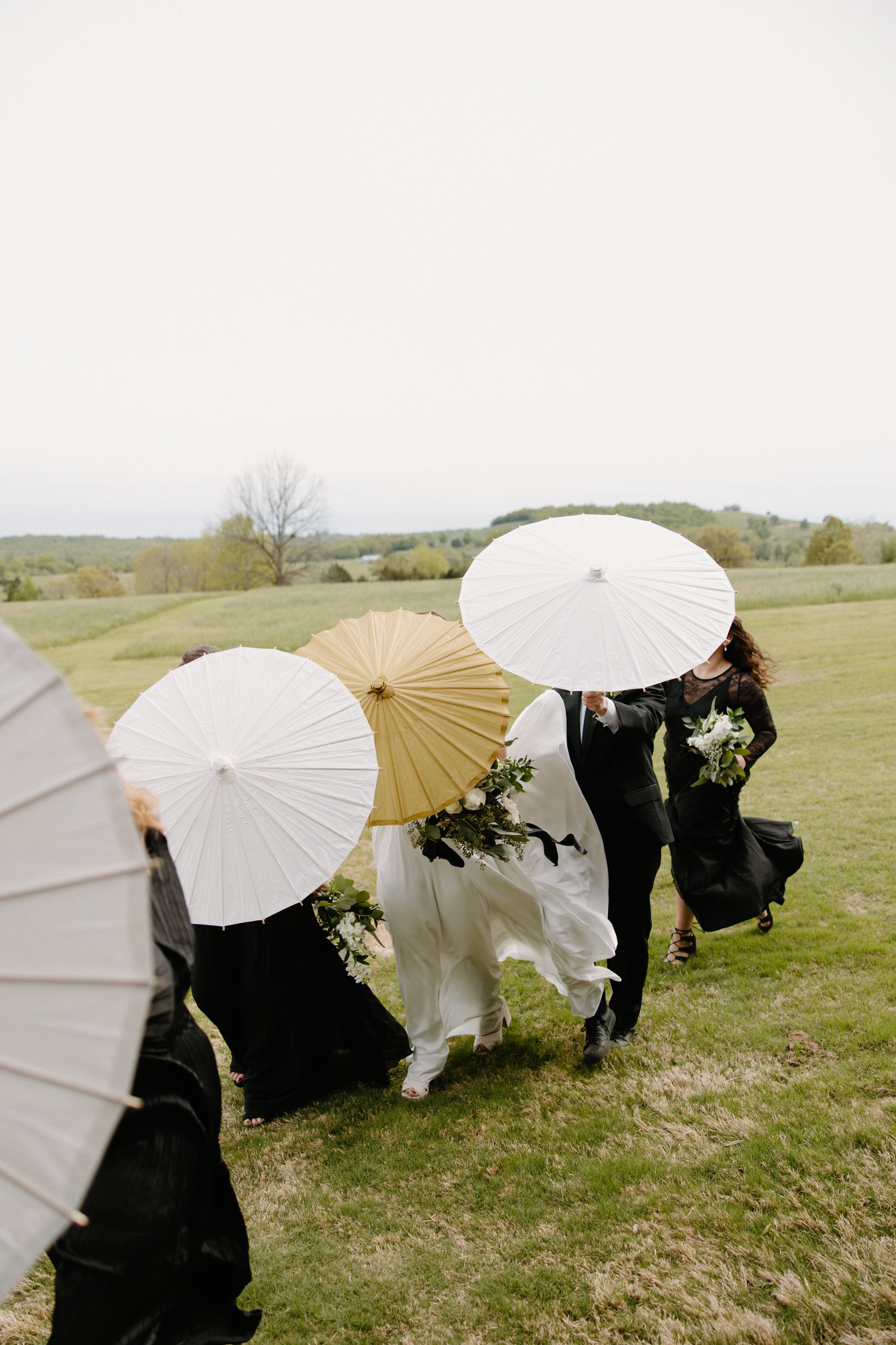 A wedding celebration outdoors with the bride and groom walking on grass while holding umbrellas to shield from rain.
