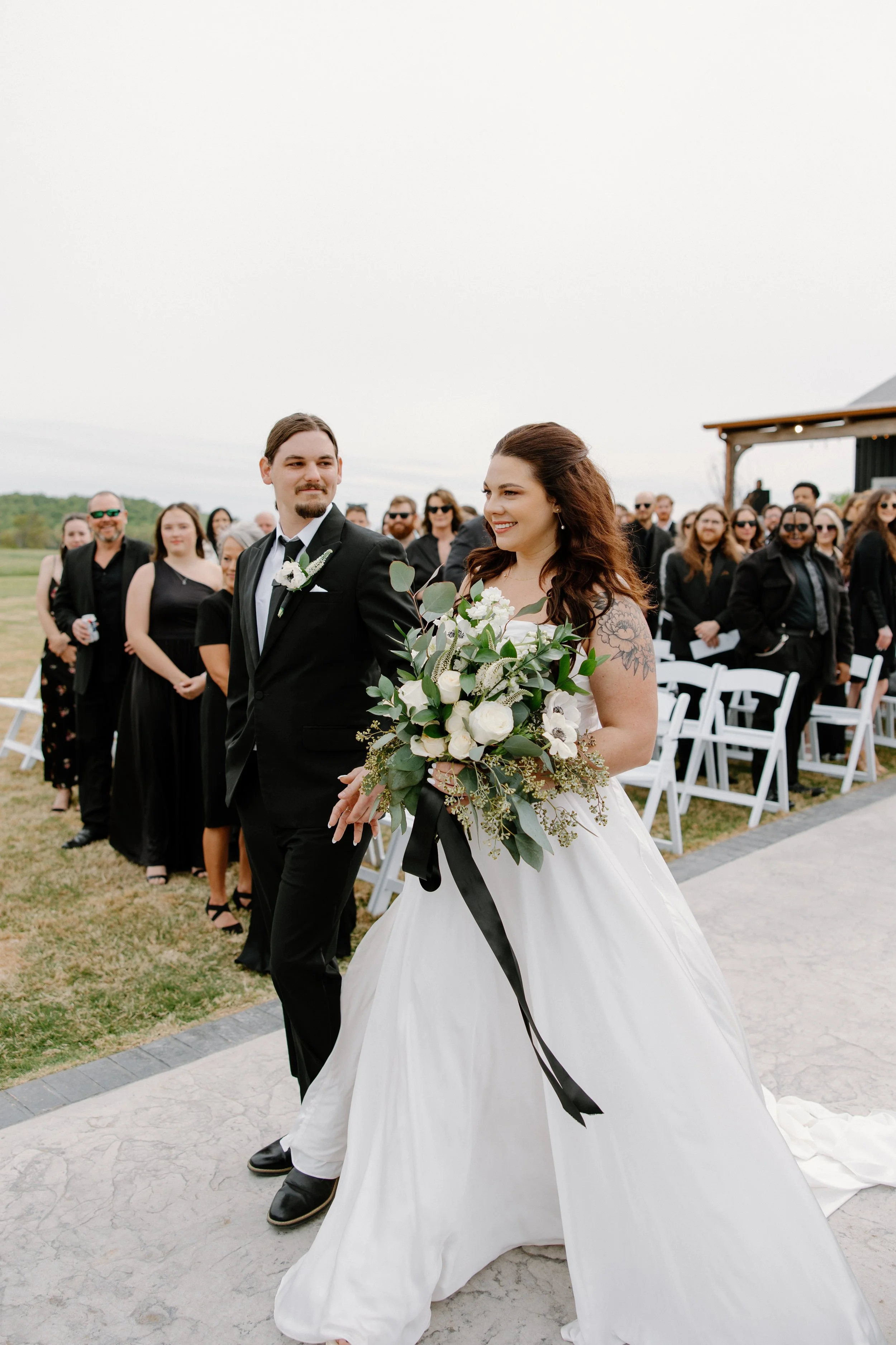 A bride and groom walking down an outdoor aisle with guests in the background at a wedding ceremony.