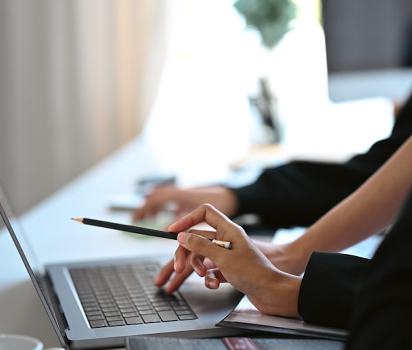 Close-up of person using a laptop and holding a pen at a desk in a well-lit office or workspace.