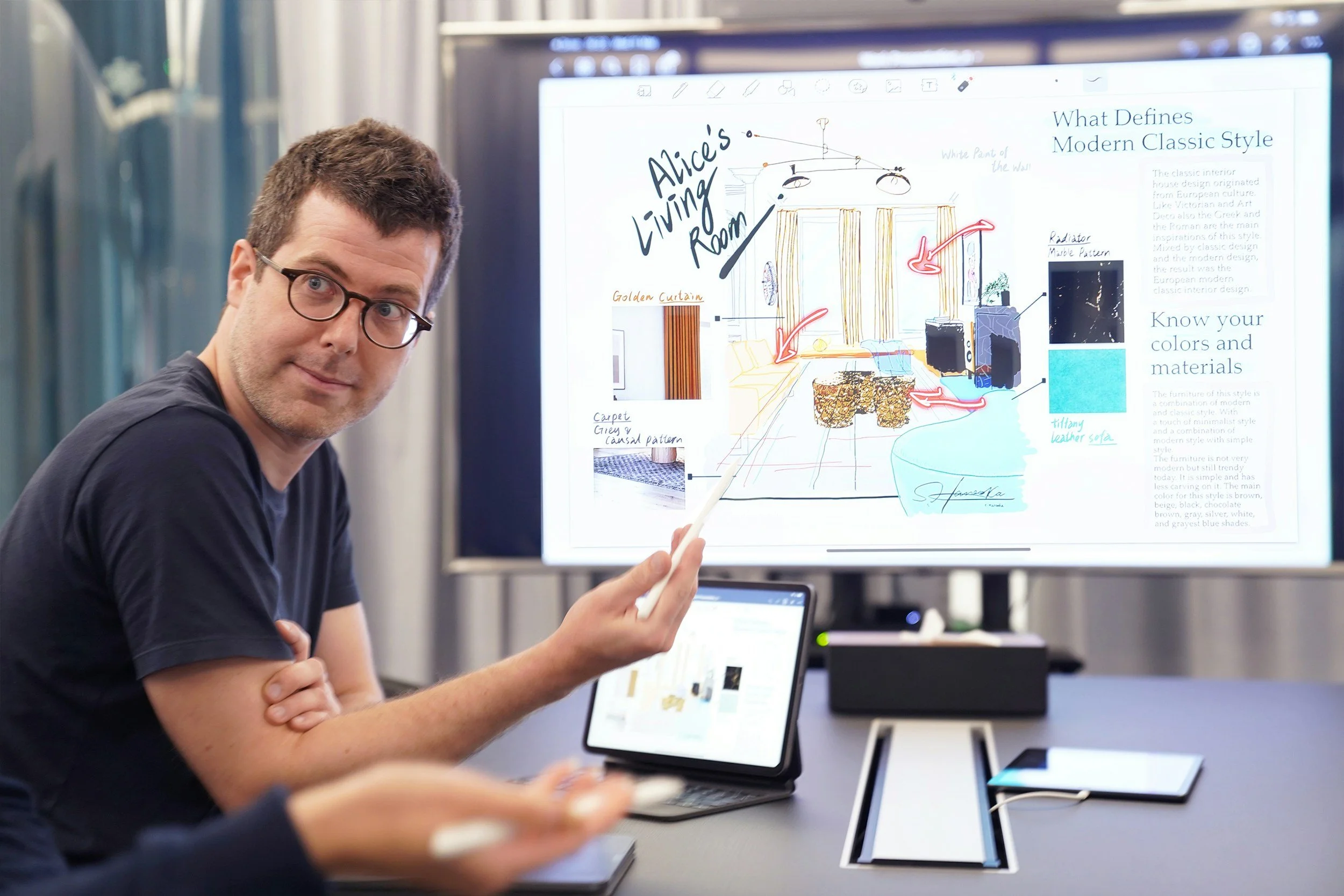 Person in glasses and black shirt presenting a digital design on a touchscreen display during a meeting in a modern conference room.