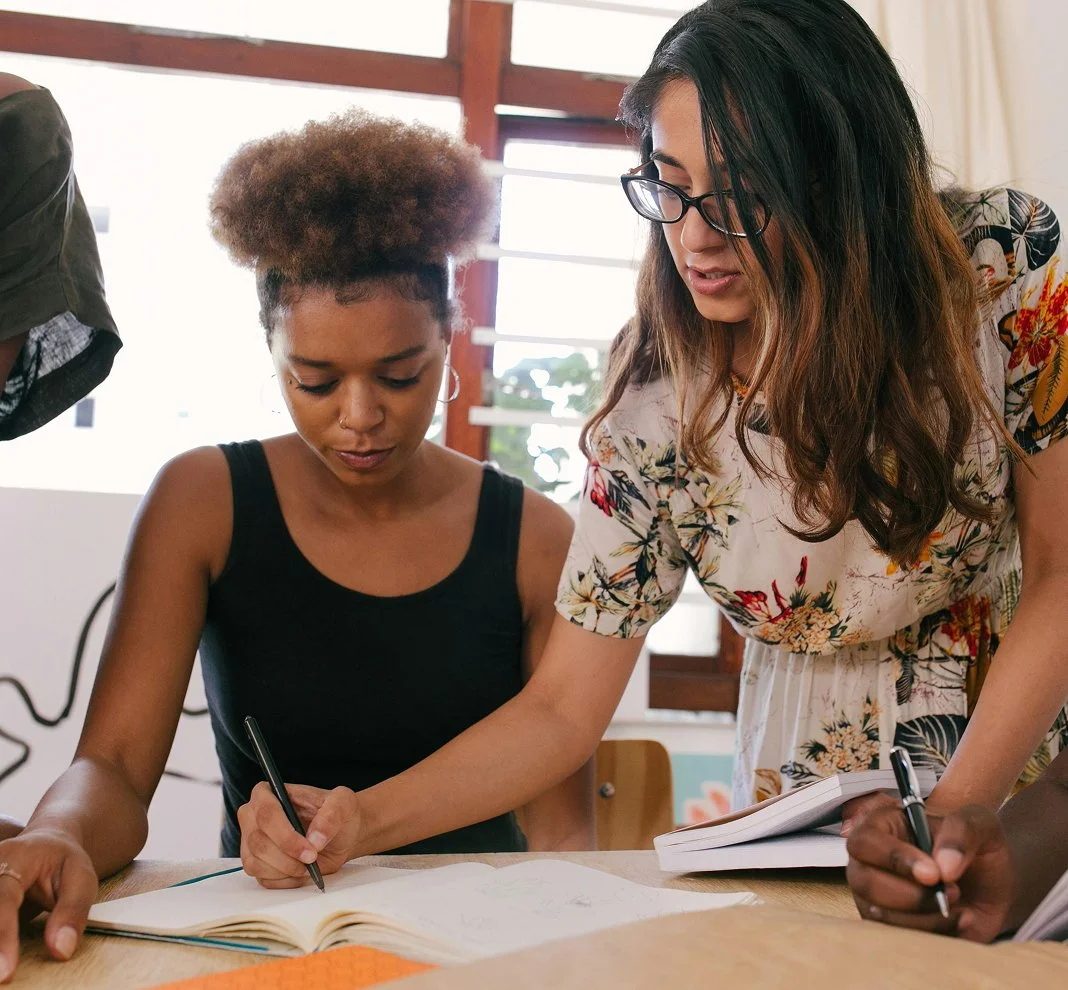 Two women working together at a table with notebooks and pens, one standing and one sitting, in a bright room with large windows.