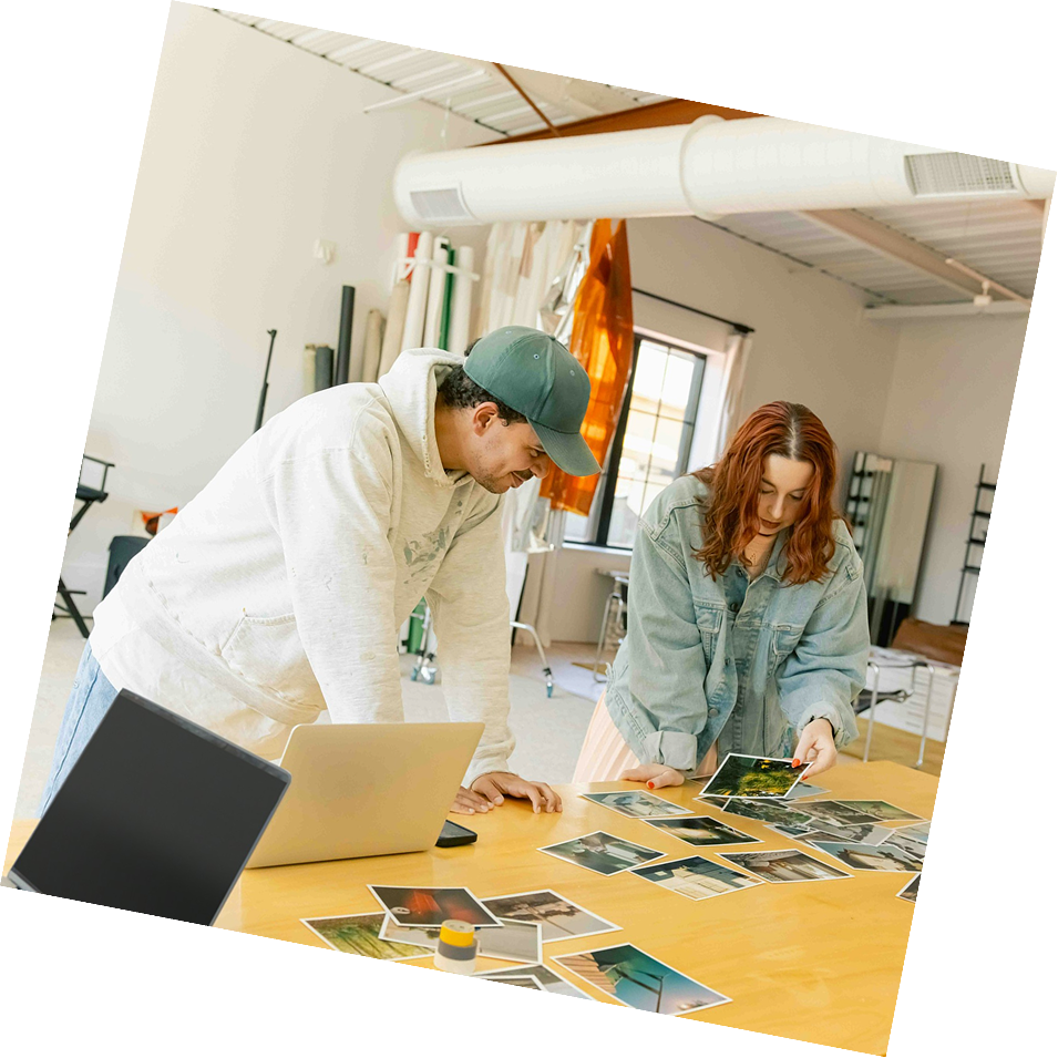 Two people, a man and a woman, look at printed photographs spread out on a table in a bright room with large windows, shelving, and equipment.