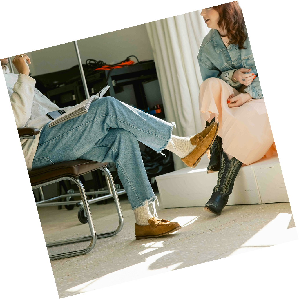 Two women talking indoors, one sitting in a chair with crossed legs and reading a book, the other sitting on a box dressed in a pink skirt and denim jacket, wearing cowboy boots.