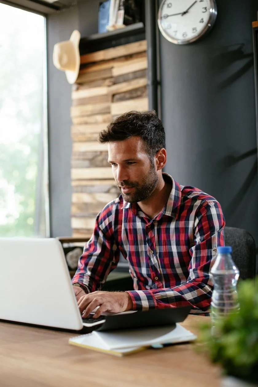 Man in plaid shirt working on a laptop at a desk with water bottle, notebook, and plant, in a room with wooden wall and clock.