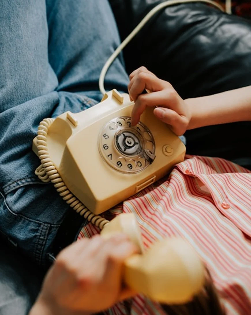 Person wearing a pink and white striped shirt and jeans holding a vintage beige rotary telephone, pressing the button to dial.
