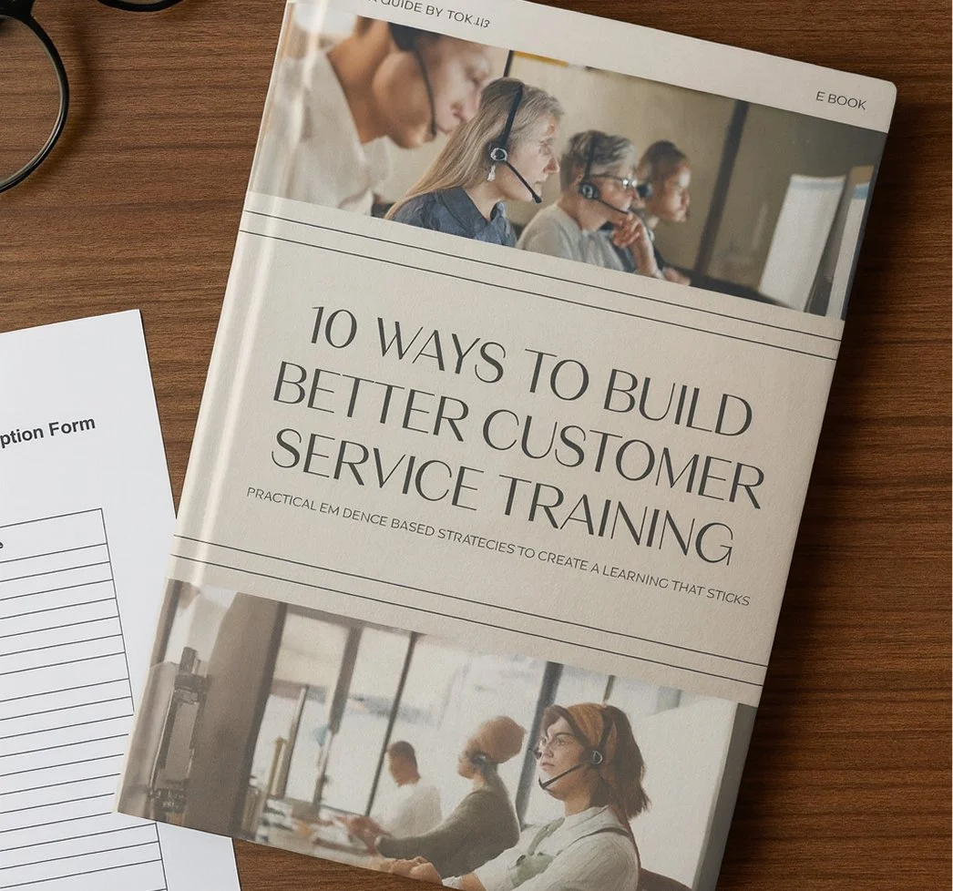 A book titled "10 Ways to Build Better Customer Service Training" on a wooden surface, with a partial view of a reception form and a black cable nearby. The book cover features images of people wearing headsets, working in customer service environments.