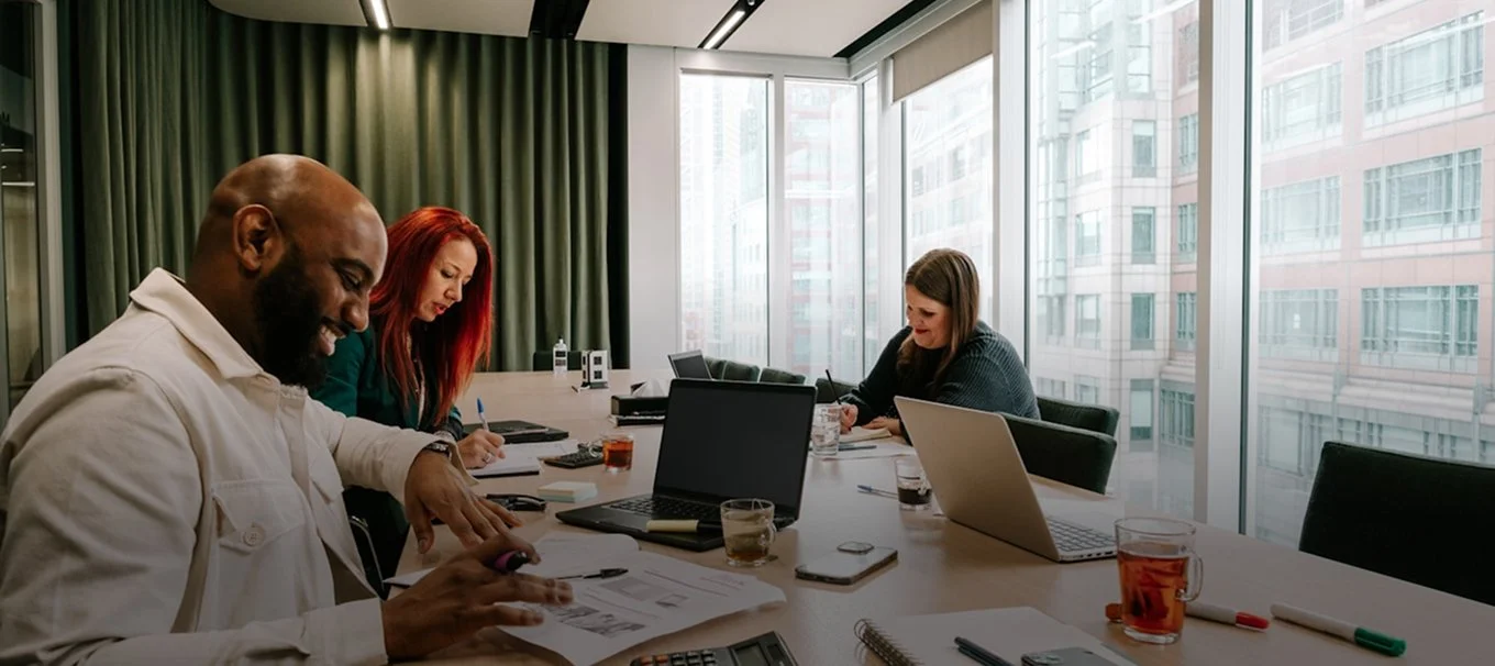 Three people sitting at a conference table in a modern office with large windows, working on laptops and reviewing papers, with drinks and office supplies on the table.