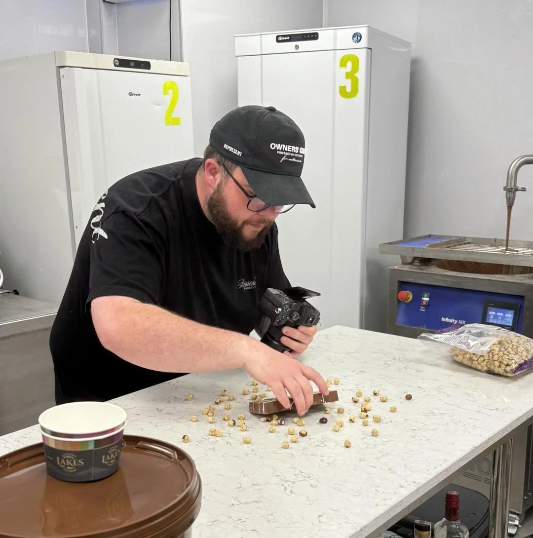 A man with glasses and a beard taking a selfie with two employees behind a chocolate shop counter. The employees are wearing white chef jackets and brown aprons with 'Castle Chocolates' embroidered on them. Decorated Christmas ornaments hang from the ceiling.